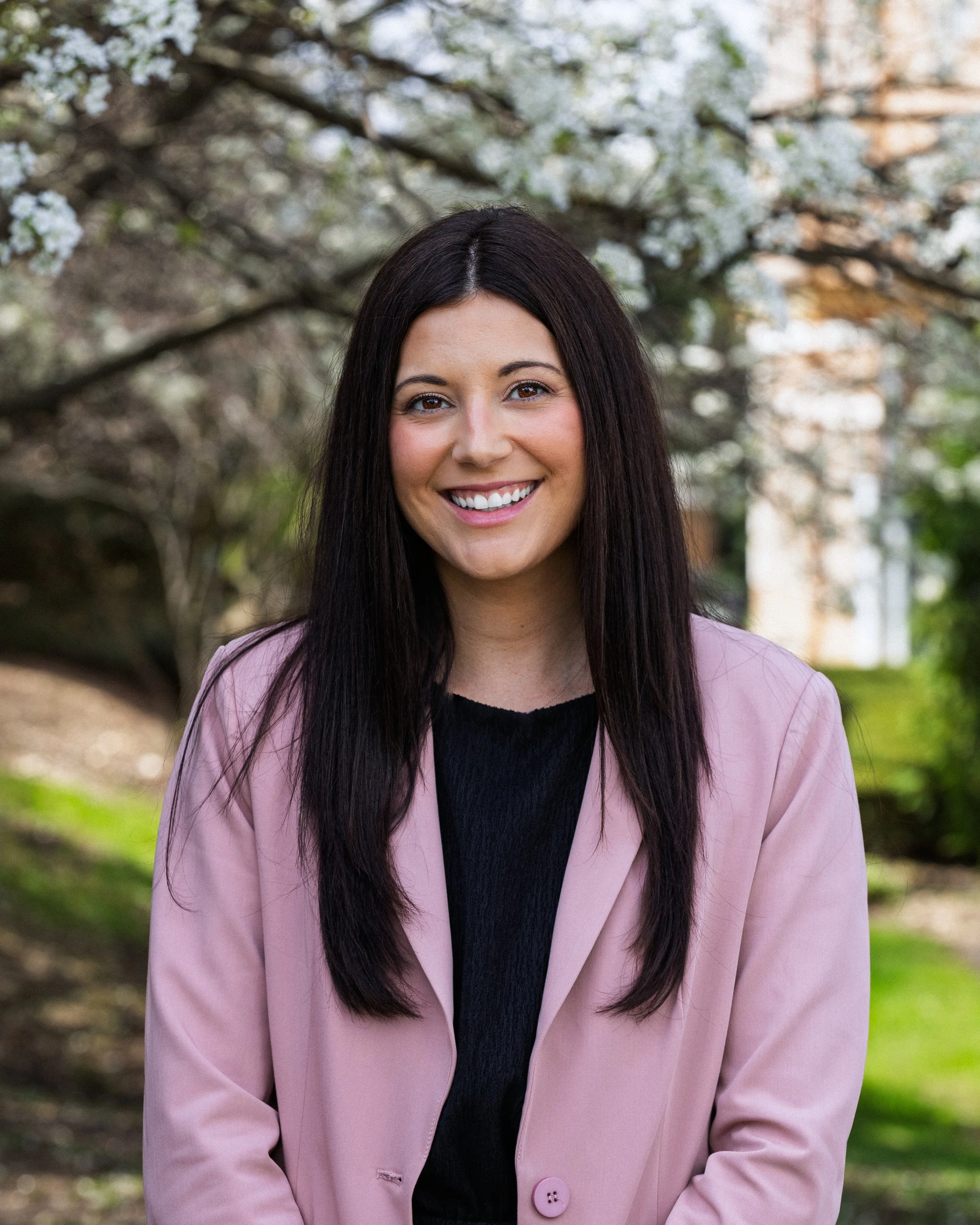 A young woman with long dark hair smiling outdoors, wearing a pink blazer over a black top. Behind her are blooming trees and greenery.