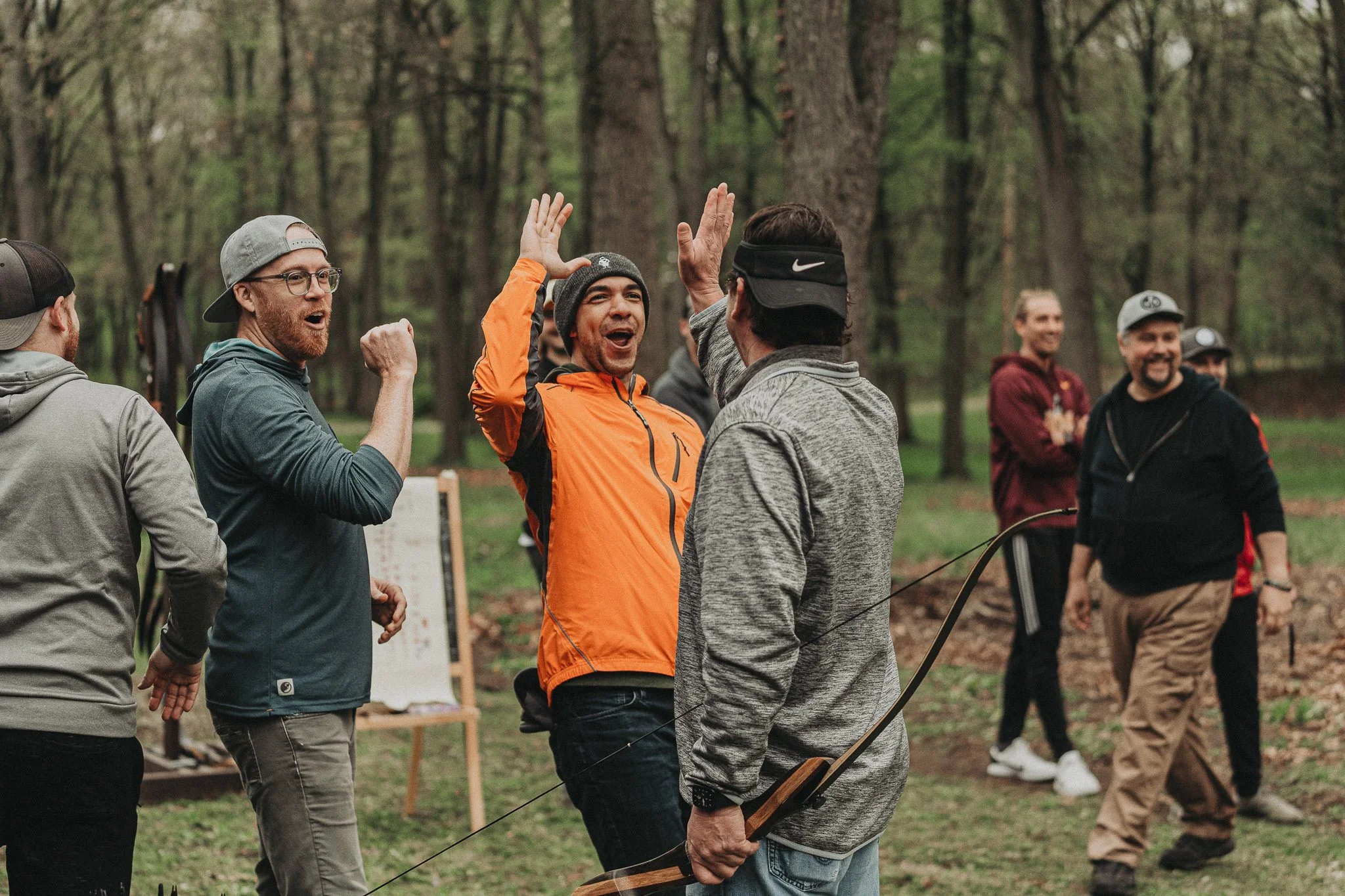 A group of people in a forest during daytime, with some high-fiving and smiling after an archery activity.