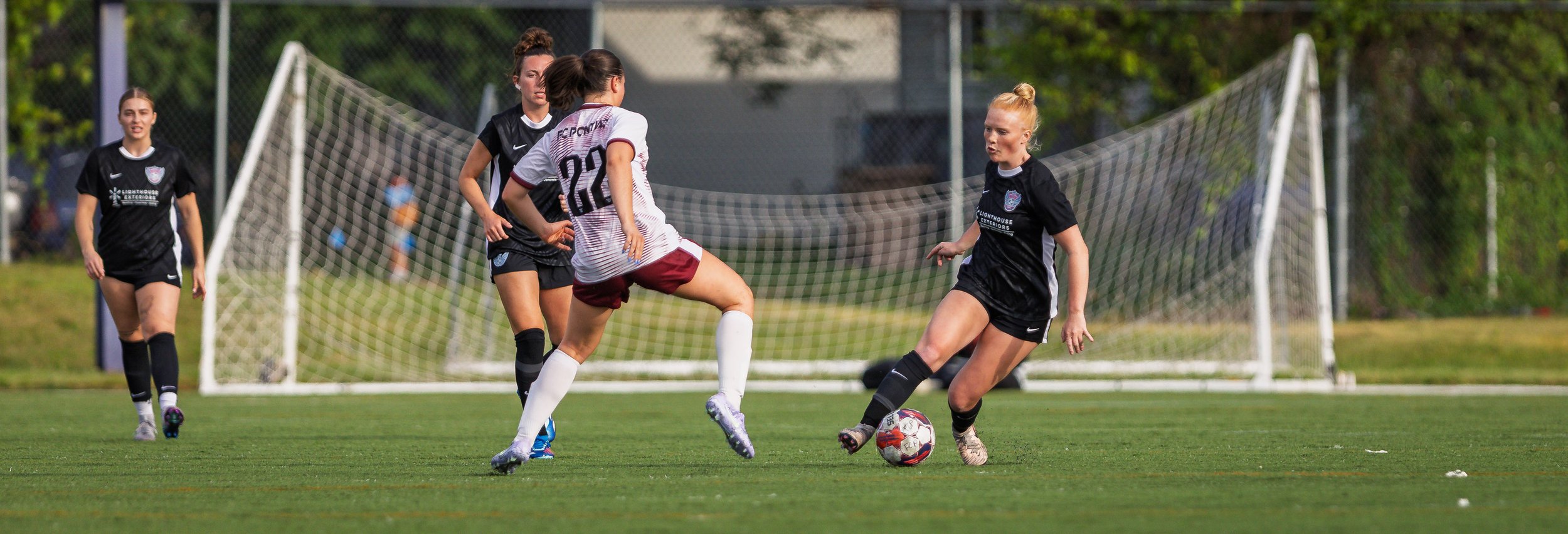 Girls playing soccer on a field, one girl in a black uniform kicking the ball while another girl in a white and maroon uniform tries to block.