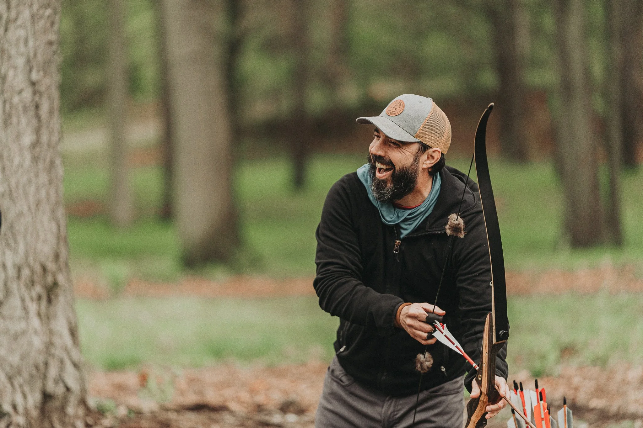 A man smiling and holding a bow and arrow outdoors in a forested area, preparing to shoot an arrow.