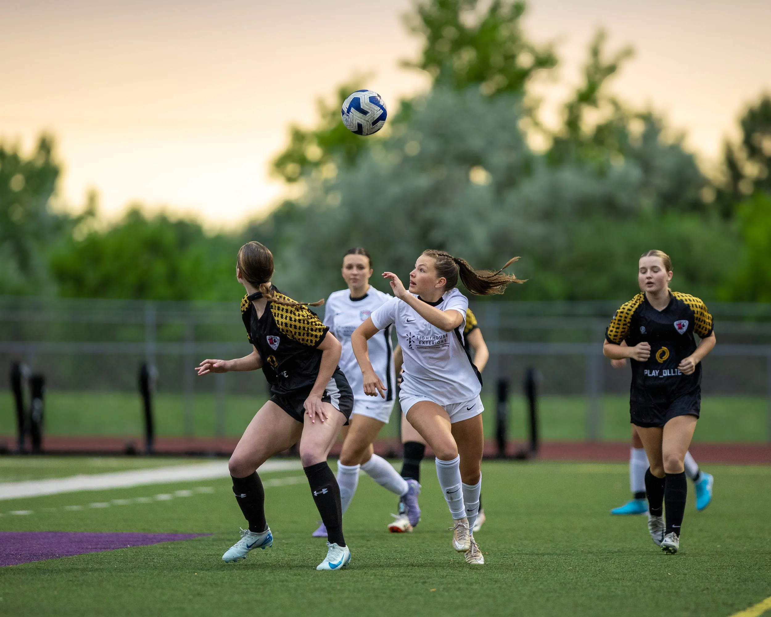 Female soccer players in black and white jerseys competing for a ball on a field during sunset.