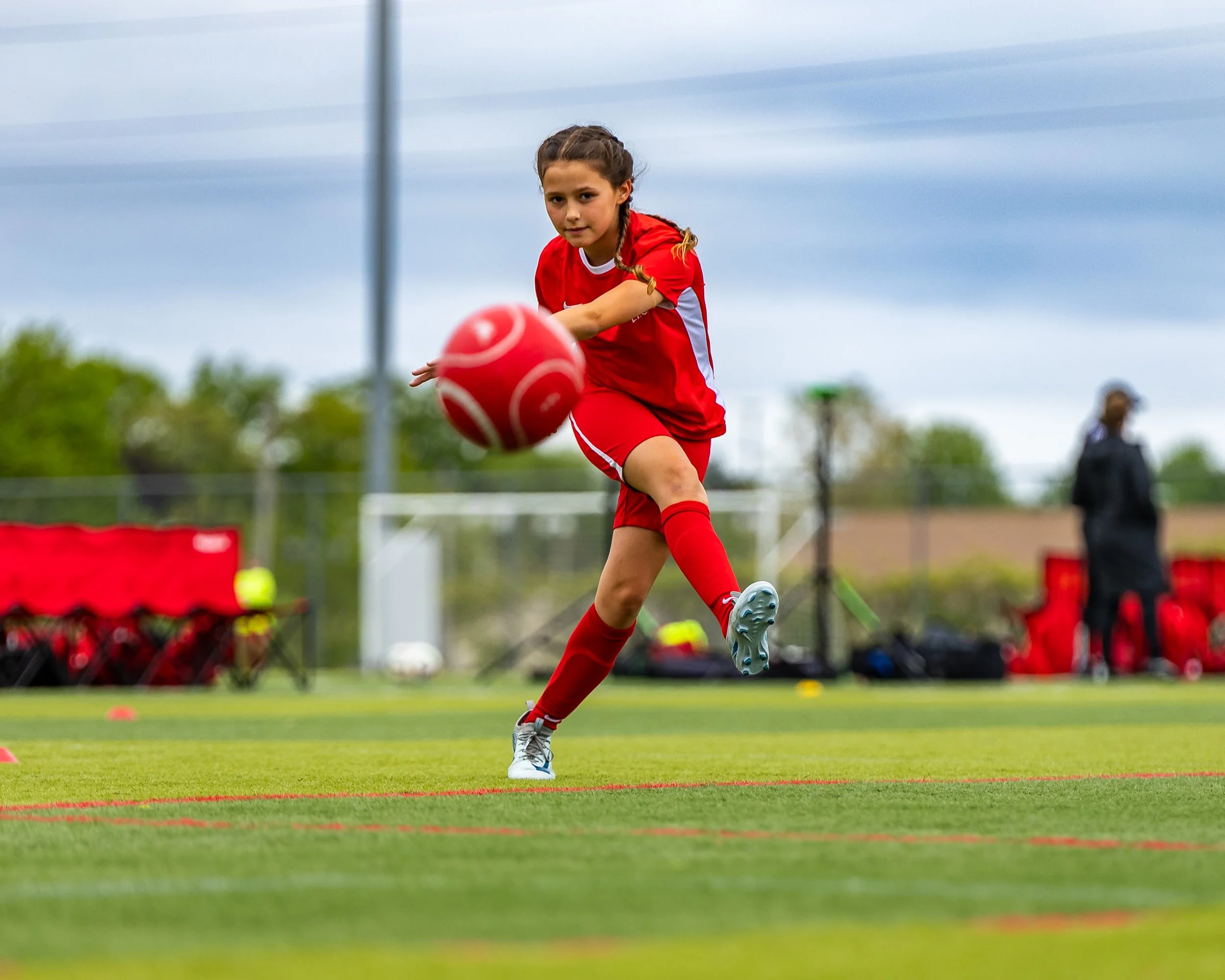 A young girl in red sports uniform kicking a red soccer ball on a grassy field