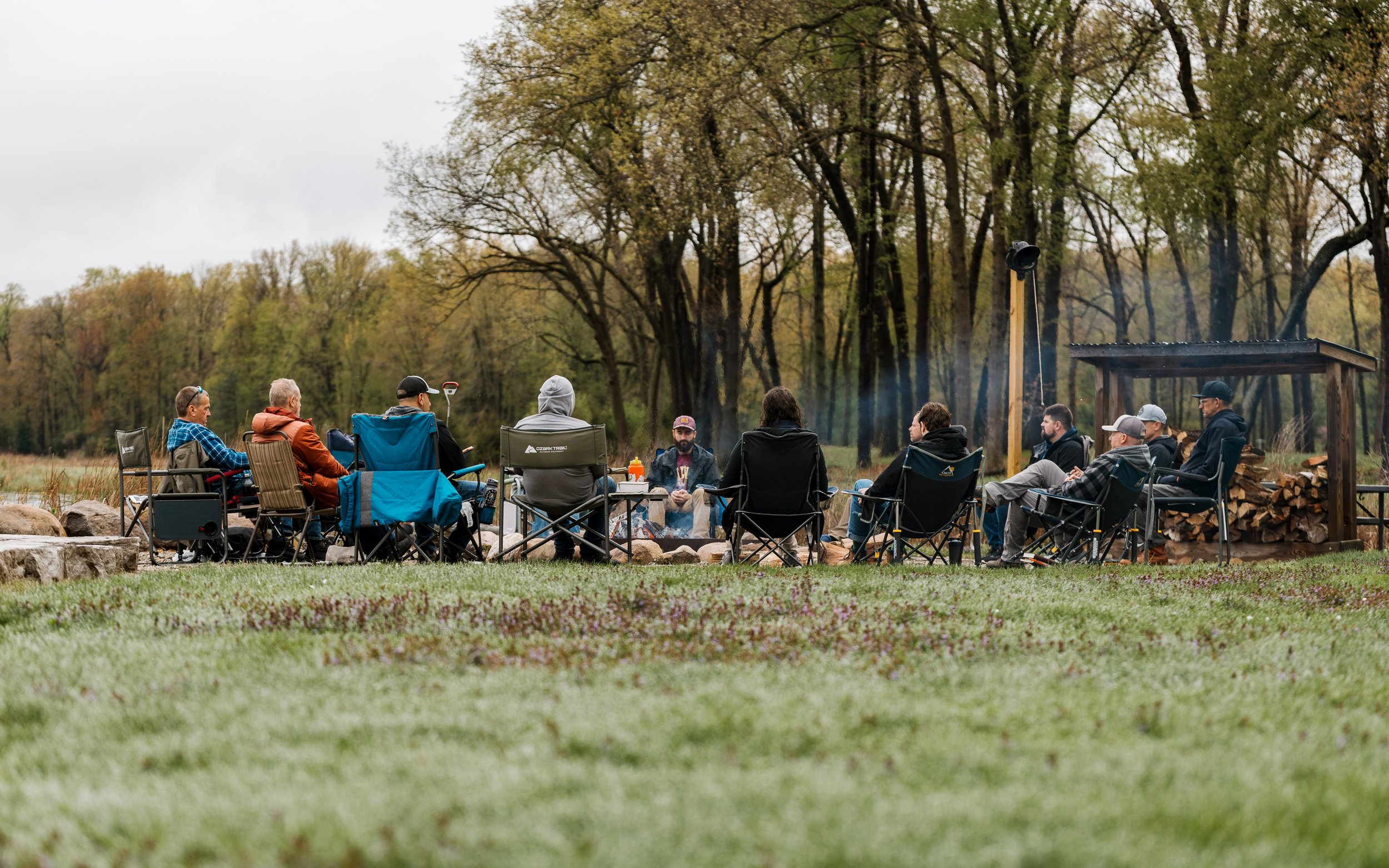 Group of people sitting around a campfire outdoors in a natural setting during daytime, with trees in the background.