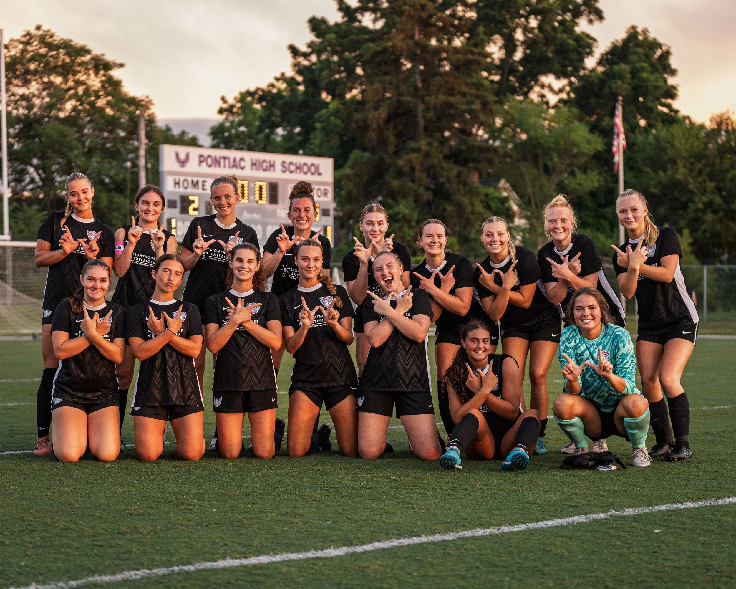 A group of female soccer players from Pontiac High School posing on the field after a game, making hand gestures and smiling.