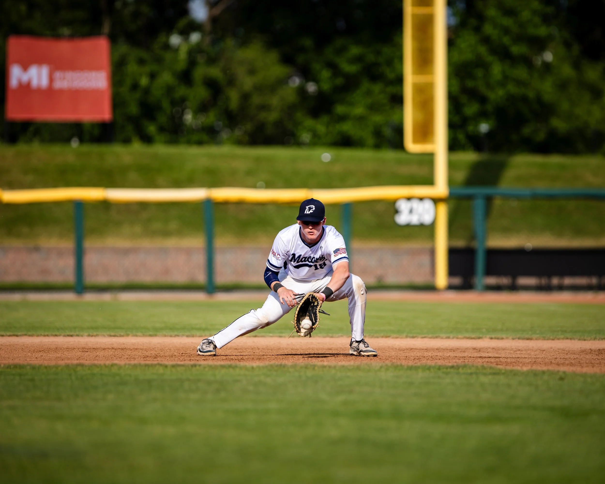 A baseball player in a white uniform with dark blue accents, wearing a cap, is fielding a ground ball on the infield. The player is crouched with a glove reaching for the ball, on a baseball field with green grass, brown dirt, and a yellow fence in t