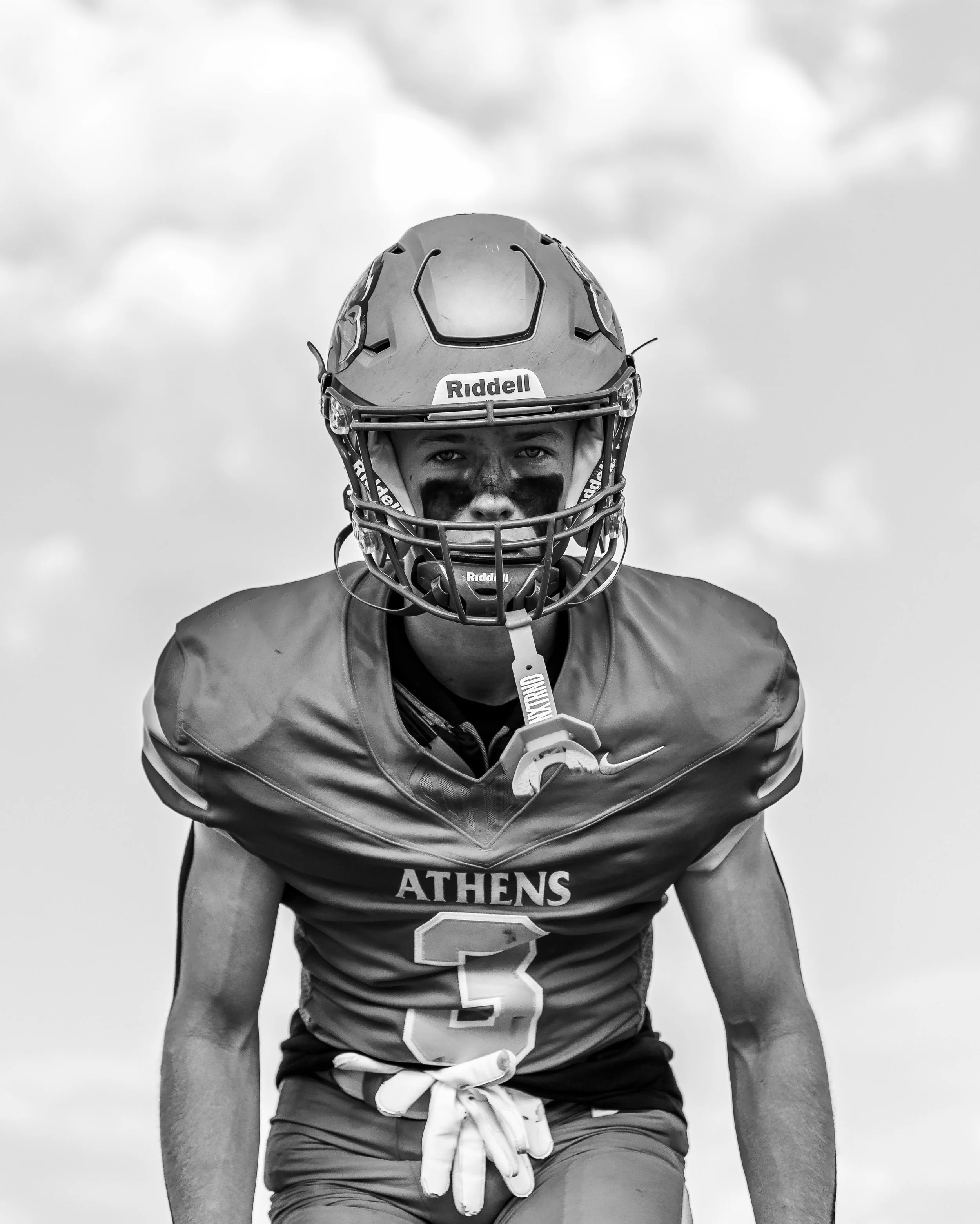 Black and white photo of a football player in uniform, wearing a helmet, shoulder pads, and gloves, standing outdoors against a cloudy sky, with a serious expression.