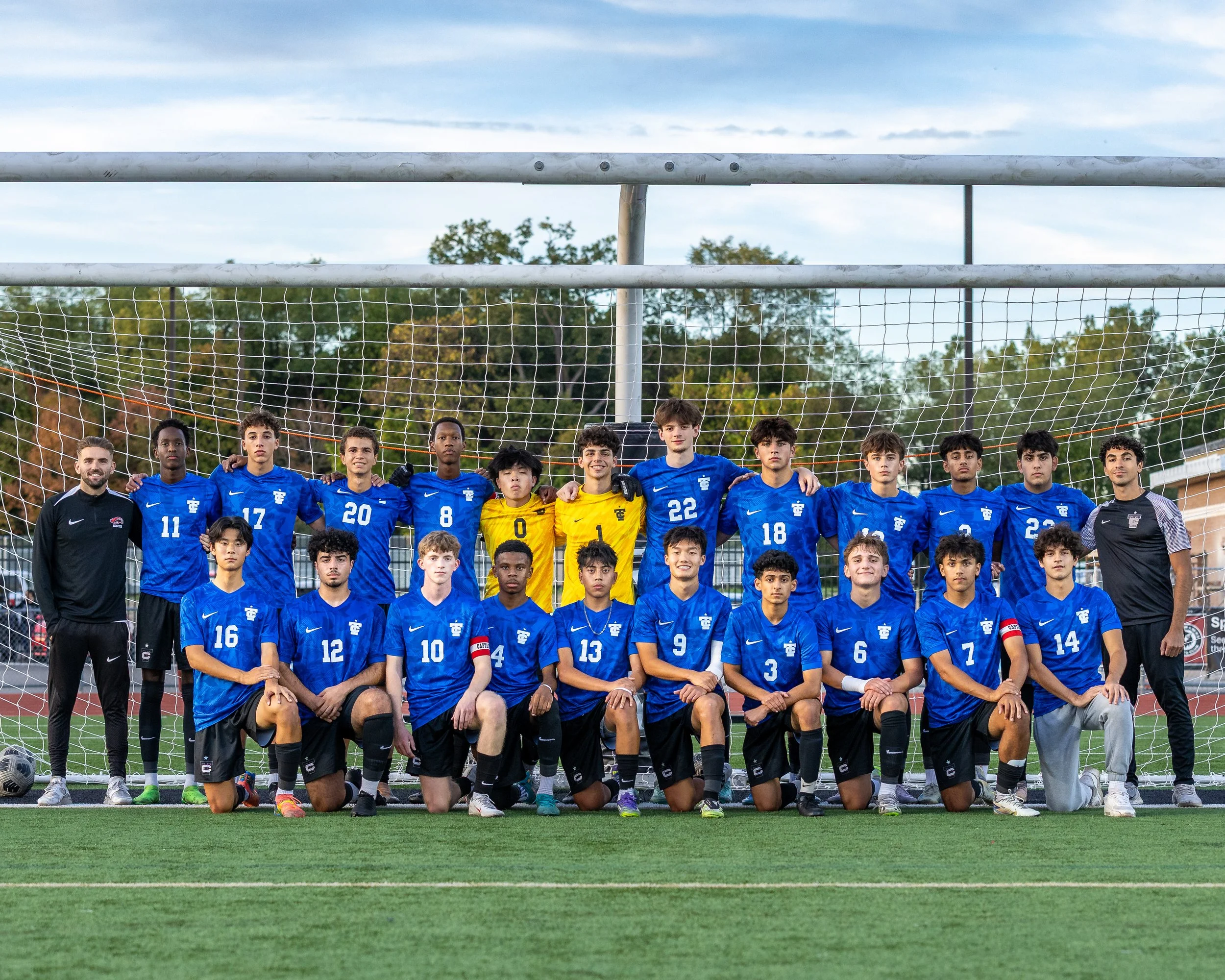 A group of soccer players and coaches on a field with goalpost and net in the background, dressed in blue jerseys with black shorts, posing for a team photo.