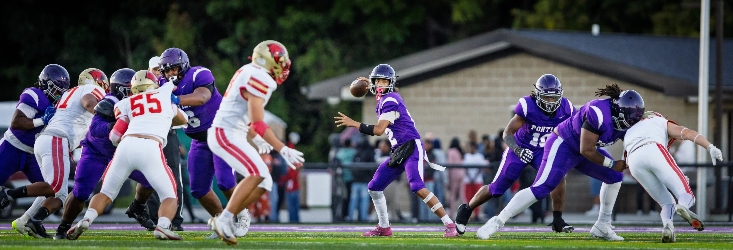 High school football game with players in purple and white uniforms on the field, quarterback in purple preparing to throw the ball, and other players engaged in blocking.