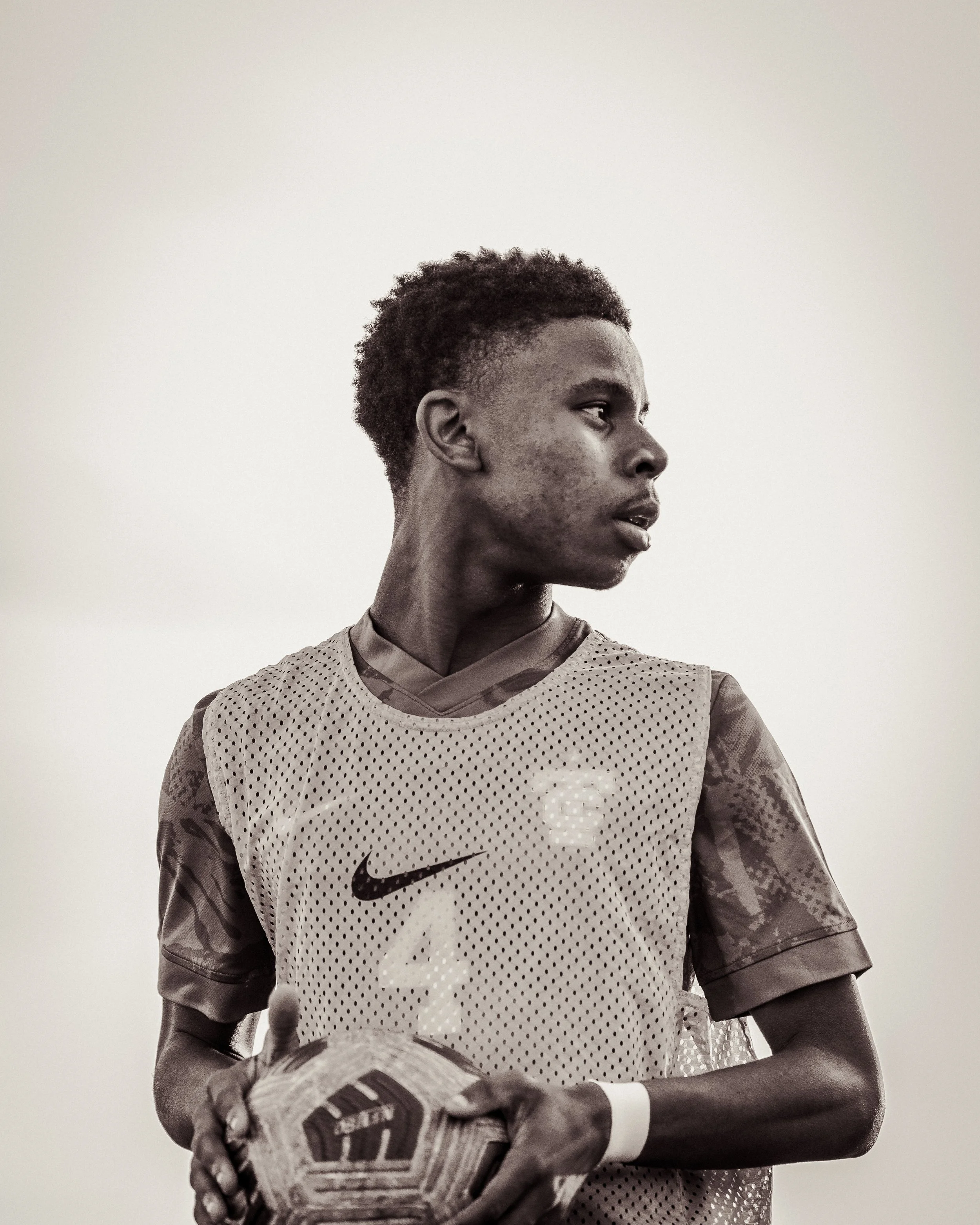 A young male soccer player with short, curly hair, wearing a beige sports vest with the Nike logo and holding a soccer ball.