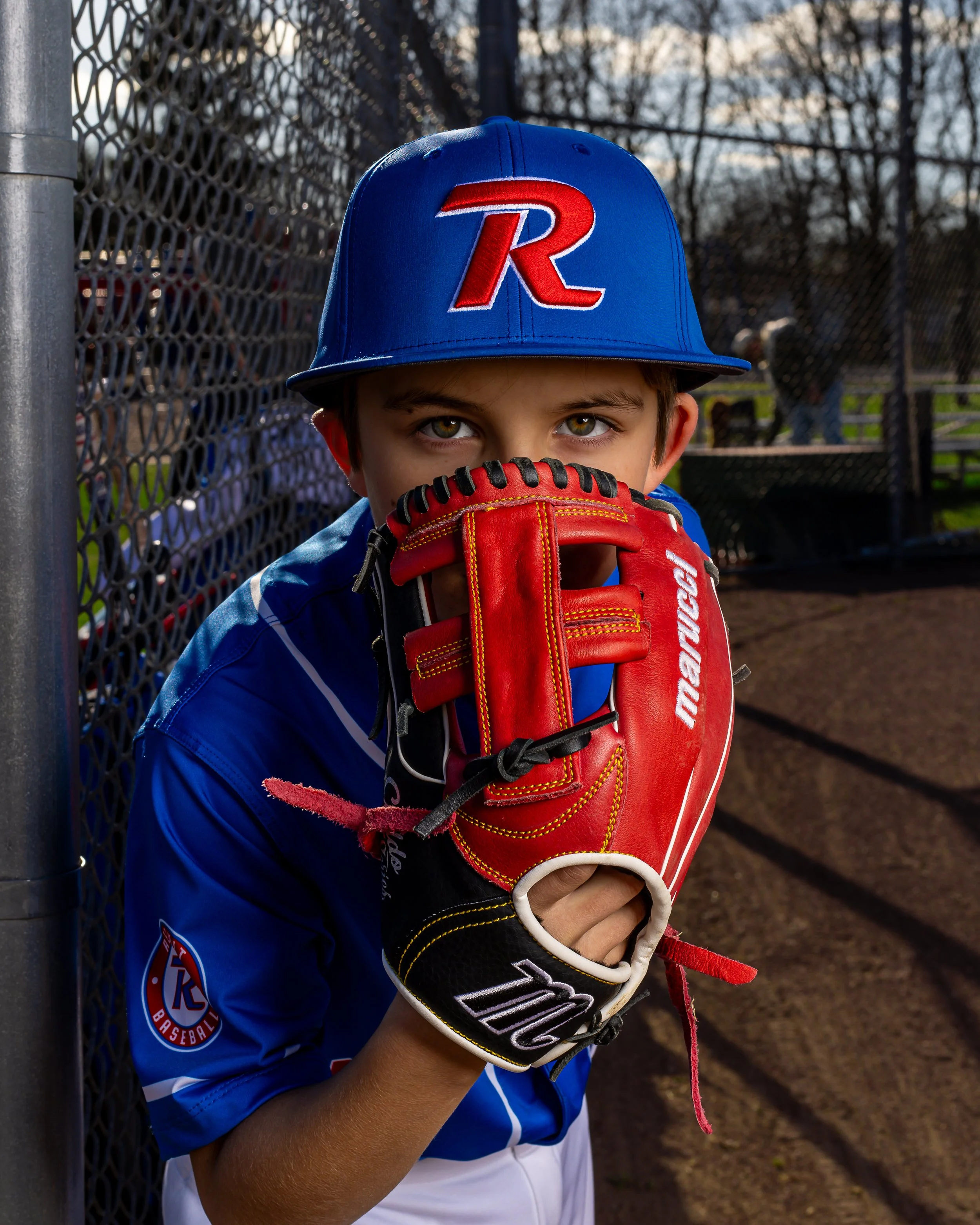 A young boy in a blue baseball uniform and blue cap with a red 'R' logo, holding a red baseball glove up to his face, standing near a chain-link fence on a baseball field.