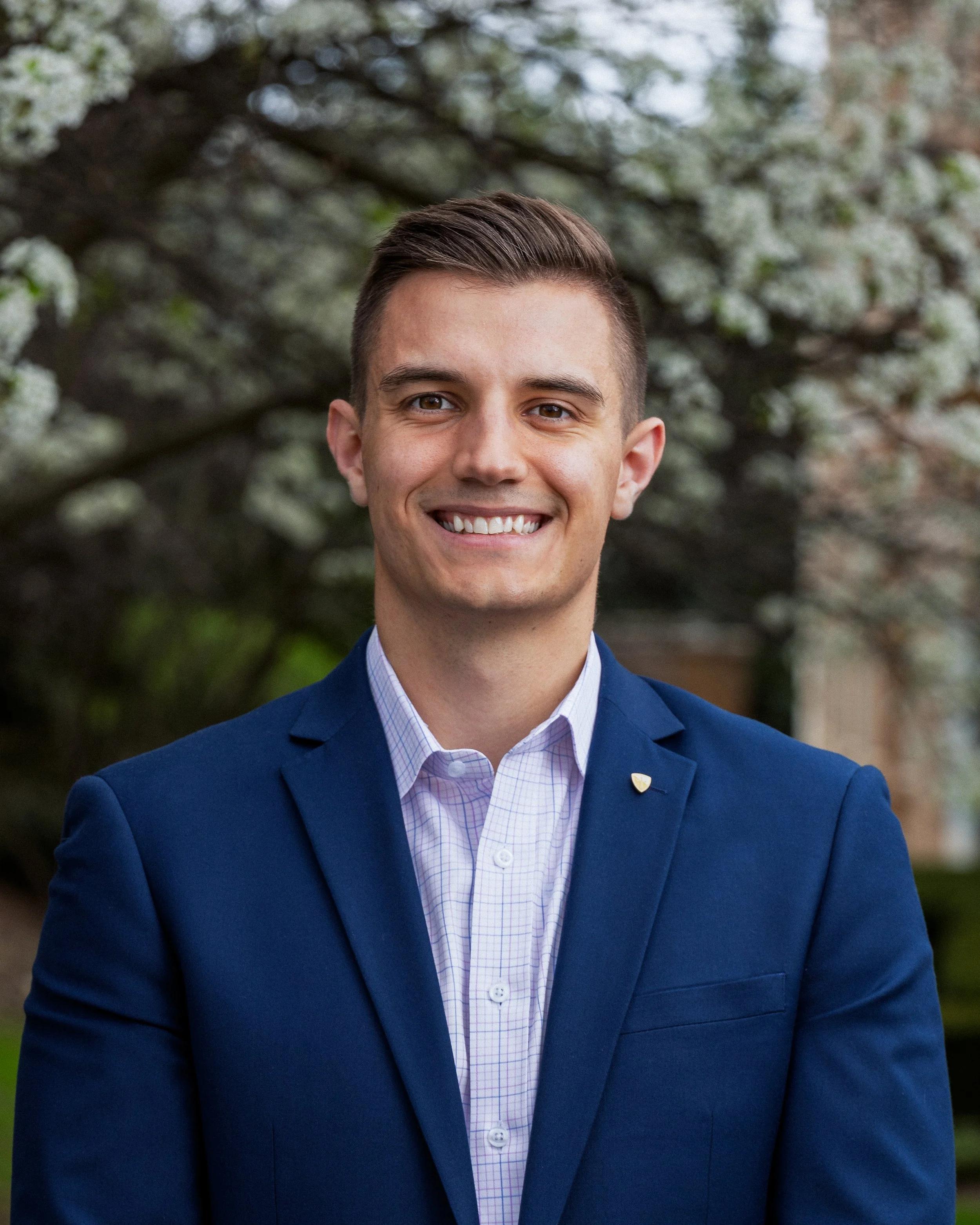 A young man with short brown hair smiling, dressed in a blue suit and light purple checkered shirt, standing outdoors with blooming trees in the background.