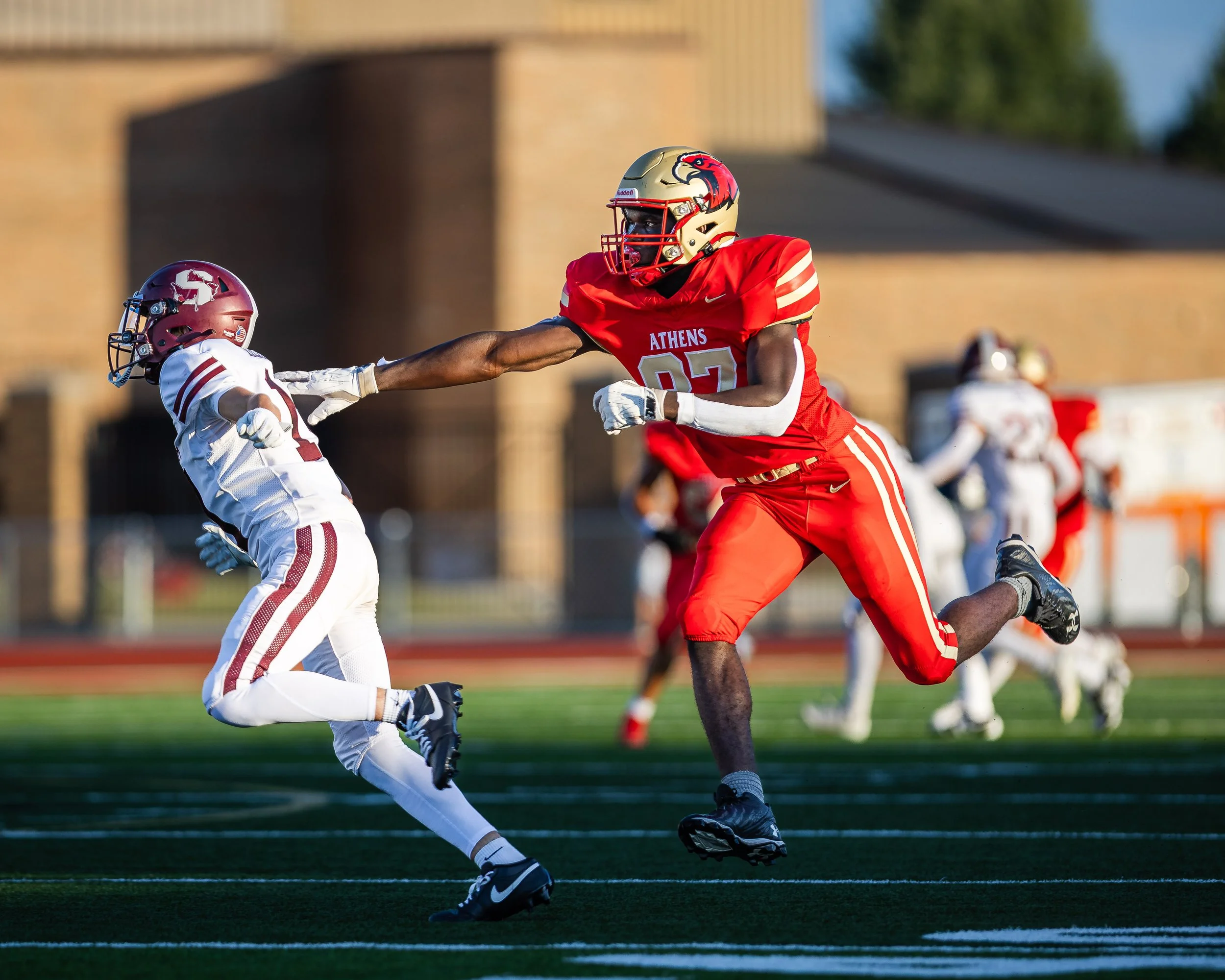 A football game with a player in a red uniform reaching out to block a player in a white uniform with a helmet, on a football field during daylight.