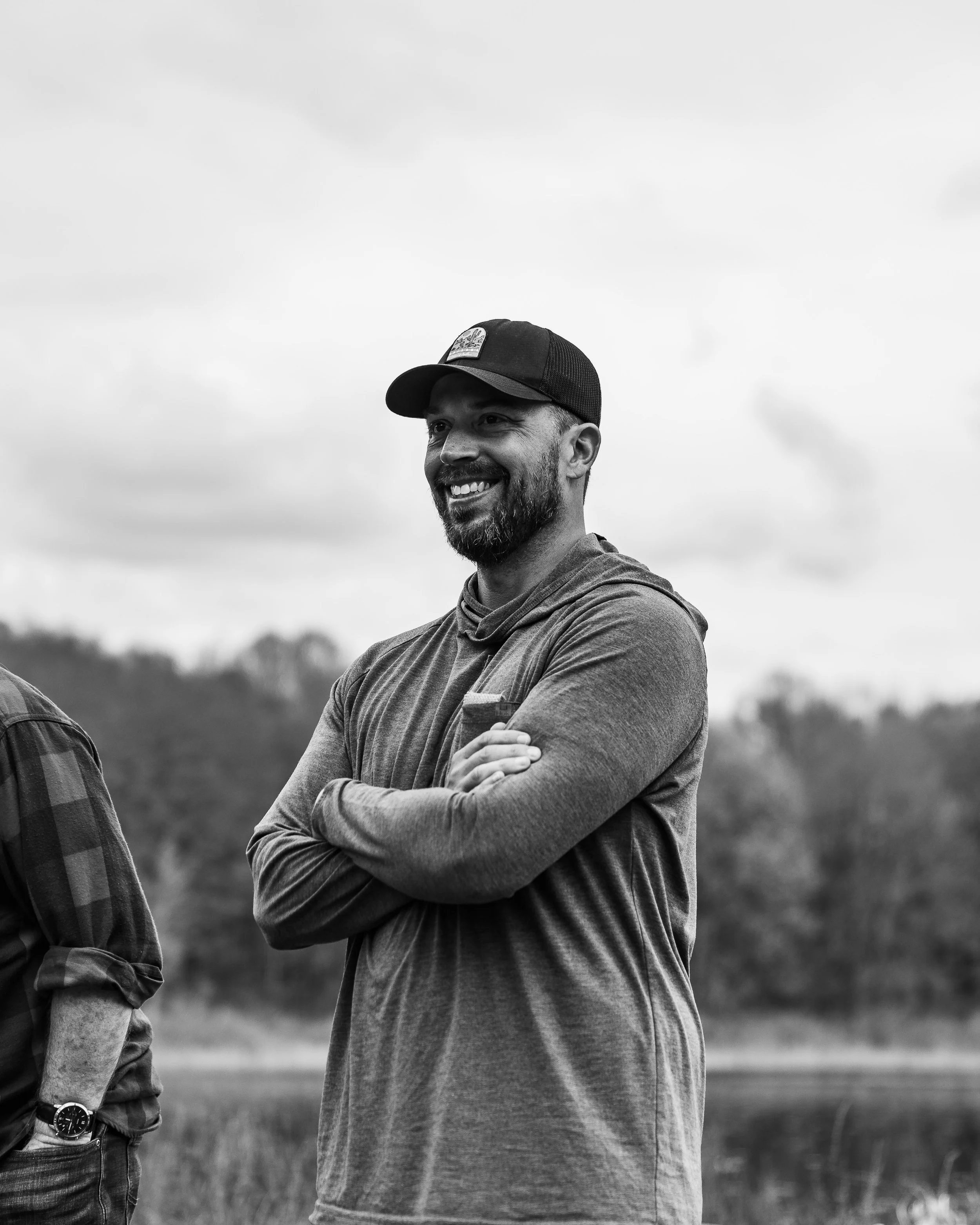 A smiling man with a beard, wearing a baseball cap and a long-sleeve shirt, standing outdoors with crossed arms.
