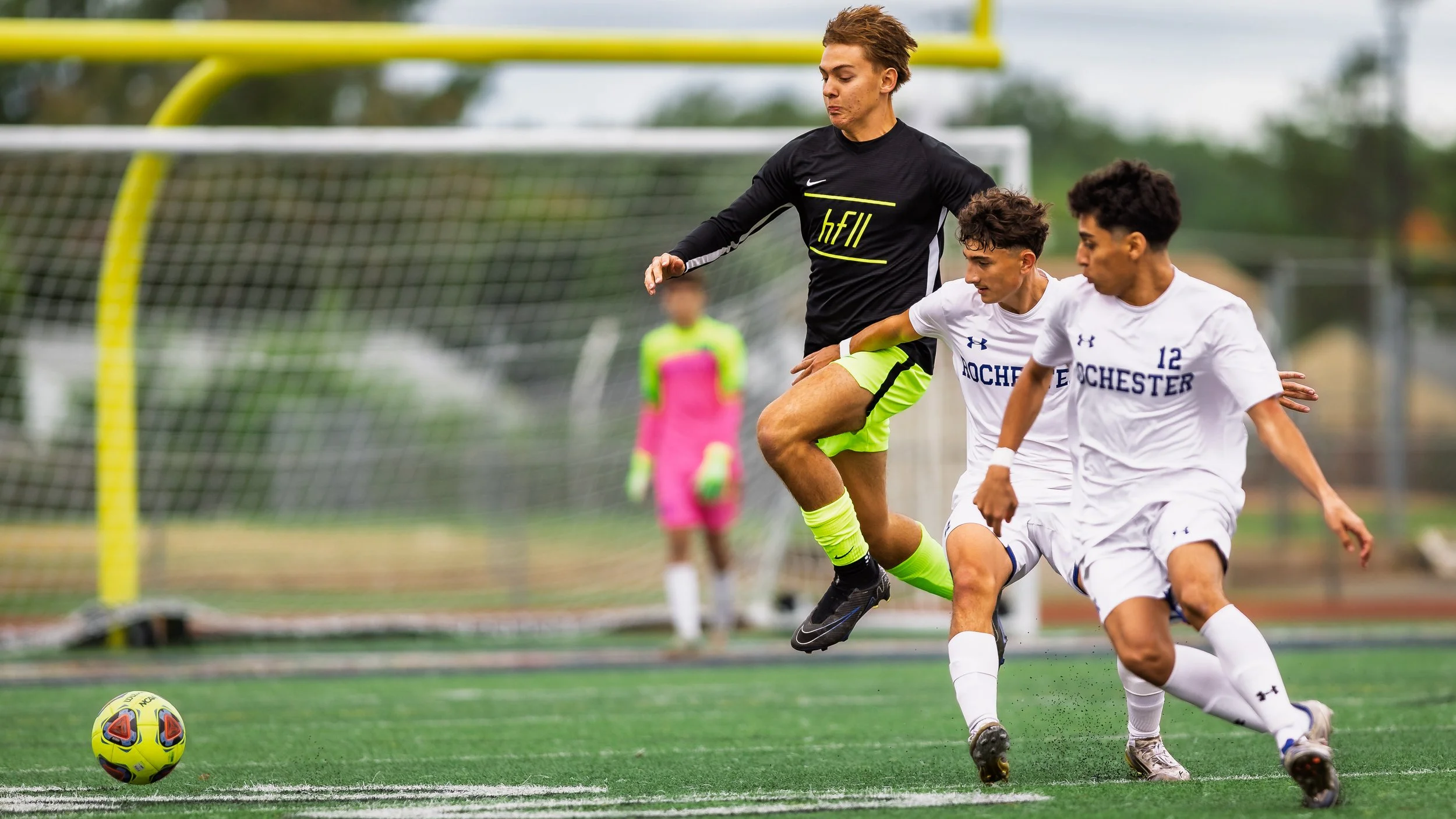 Soccer players in white and black uniforms competing for the ball on a field, with a goalkeeper in pink in the background.