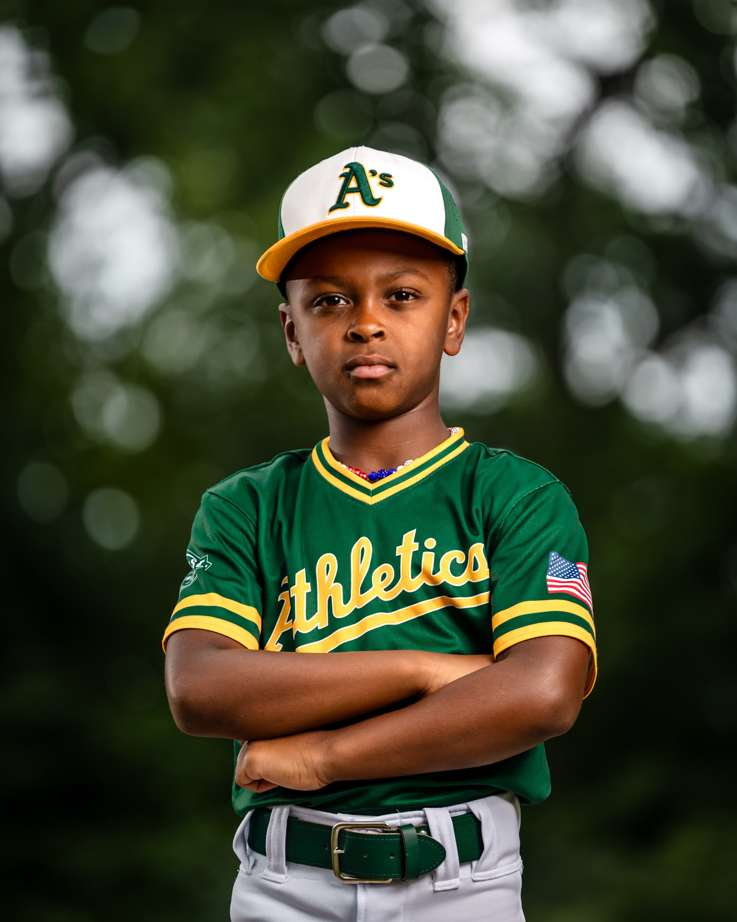 Young boy in a green Oakland Athletics baseball uniform with a cap, standing outdoors with arms crossed and serious expression.