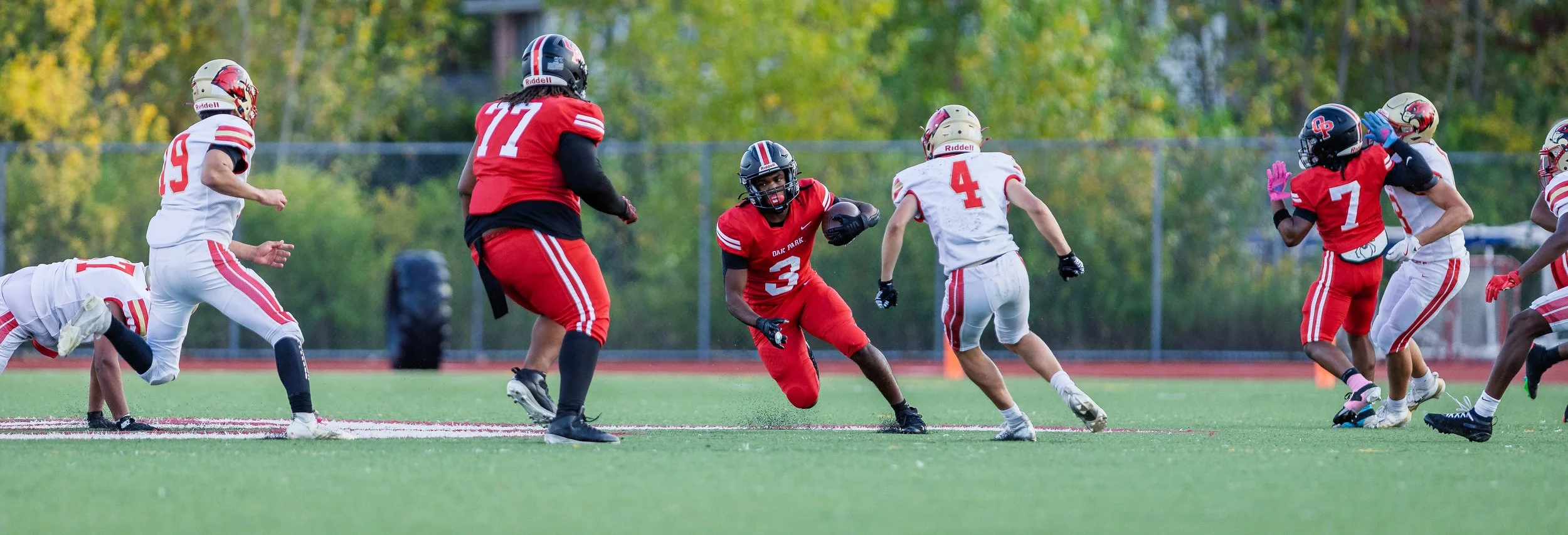 Youth football game with players in red and white jerseys on a grassy field, some players running and others tackling, with trees in the background.