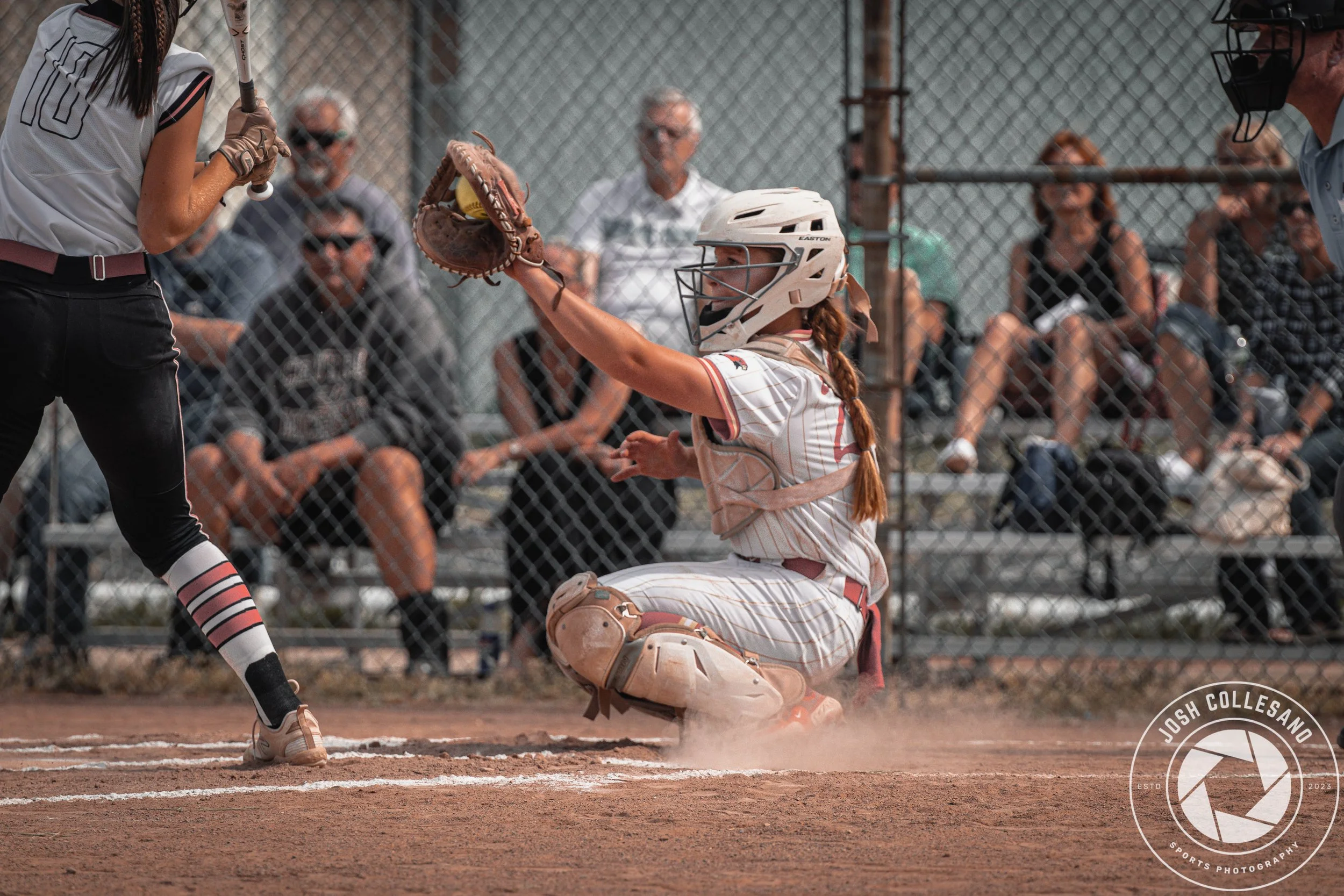 A female softball catcher wearing a helmet and baseball uniform, squatting behind home plate, catching a pitch. An umpire is standing nearby, and an opposing player is at bat. Spectators sit behind a chain-link fence in the background.