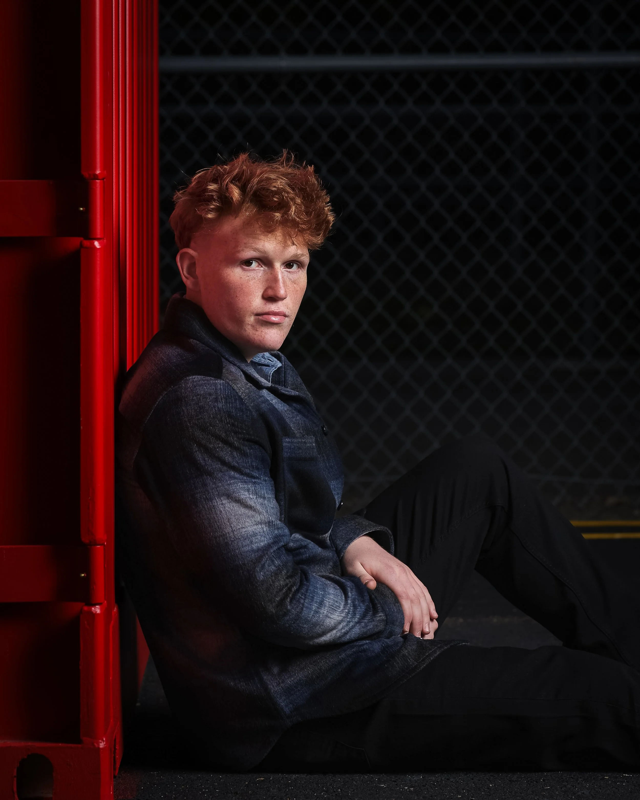 A young red-haired man sitting on the ground against a black chain-link fence, wearing a dark jacket and pants, looking at the camera with a serious expression.