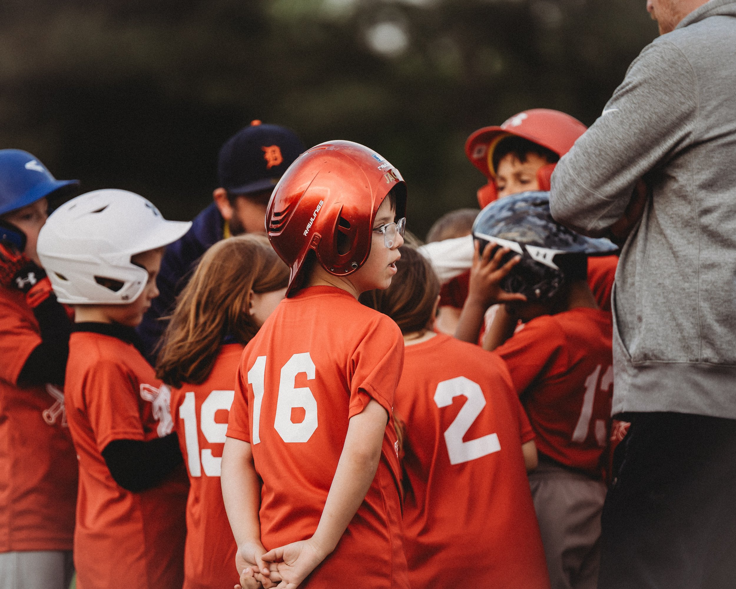 Youth baseball team gathered around coach during huddle, with players wearing red jerseys and helmets numbered 16, 15, 2, and others.