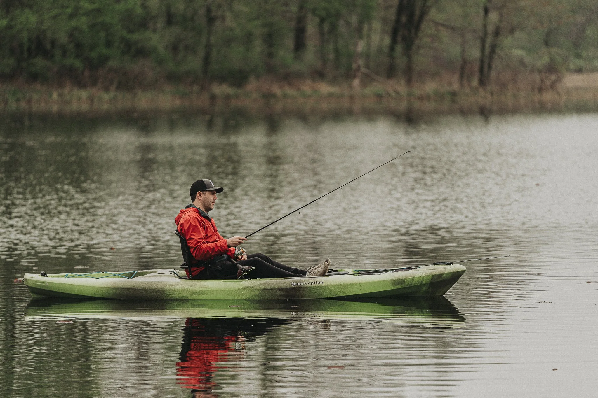 A man in a red jacket and black cap fishing from a green kayak on a calm lake, with trees in the background.