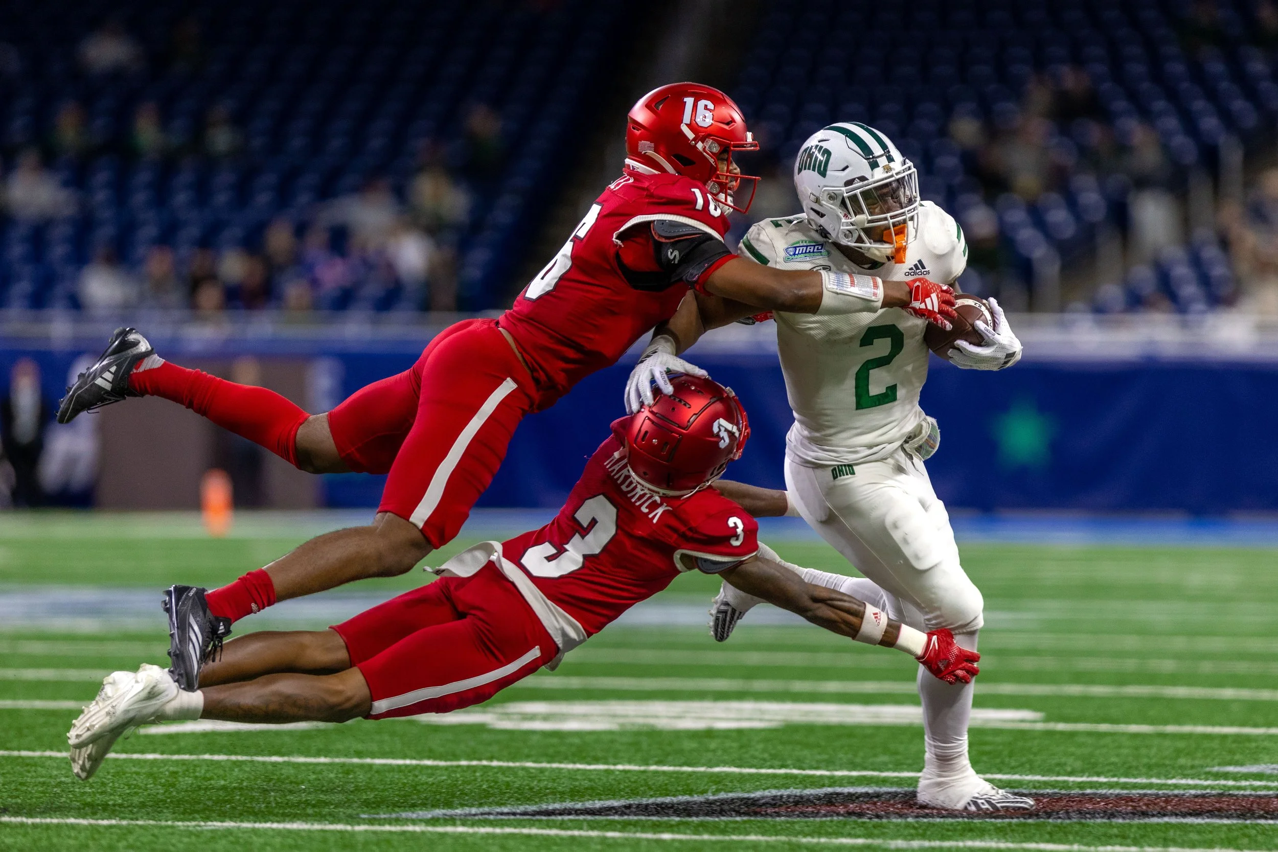 Two football players in red uniforms tackle a player in a white uniform with the number 2, who is running with the ball in an indoor stadium.