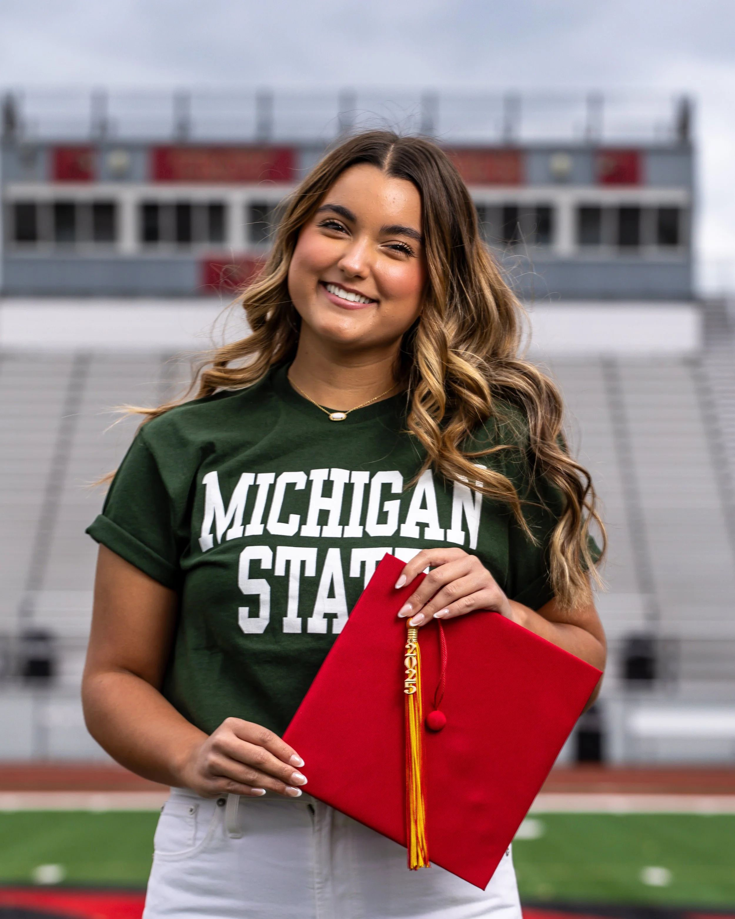 A young woman with long wavy hair smiling at the camera, wearing a green 'Michigan State' t-shirt, holding a red graduation cap with a yellow and red tassel, standing in a stadium.