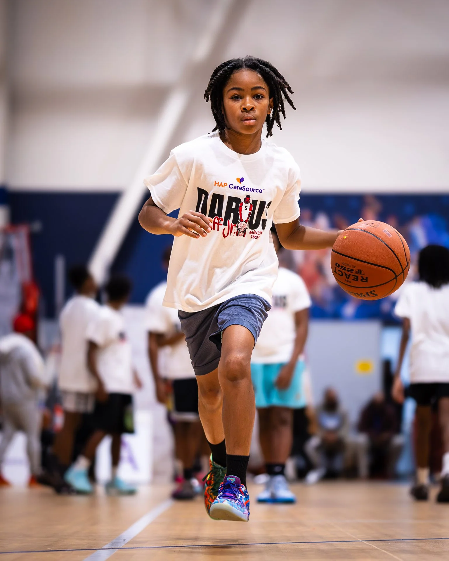 Young girl dribbling a basketball on an indoor court with other children and adults in the background.