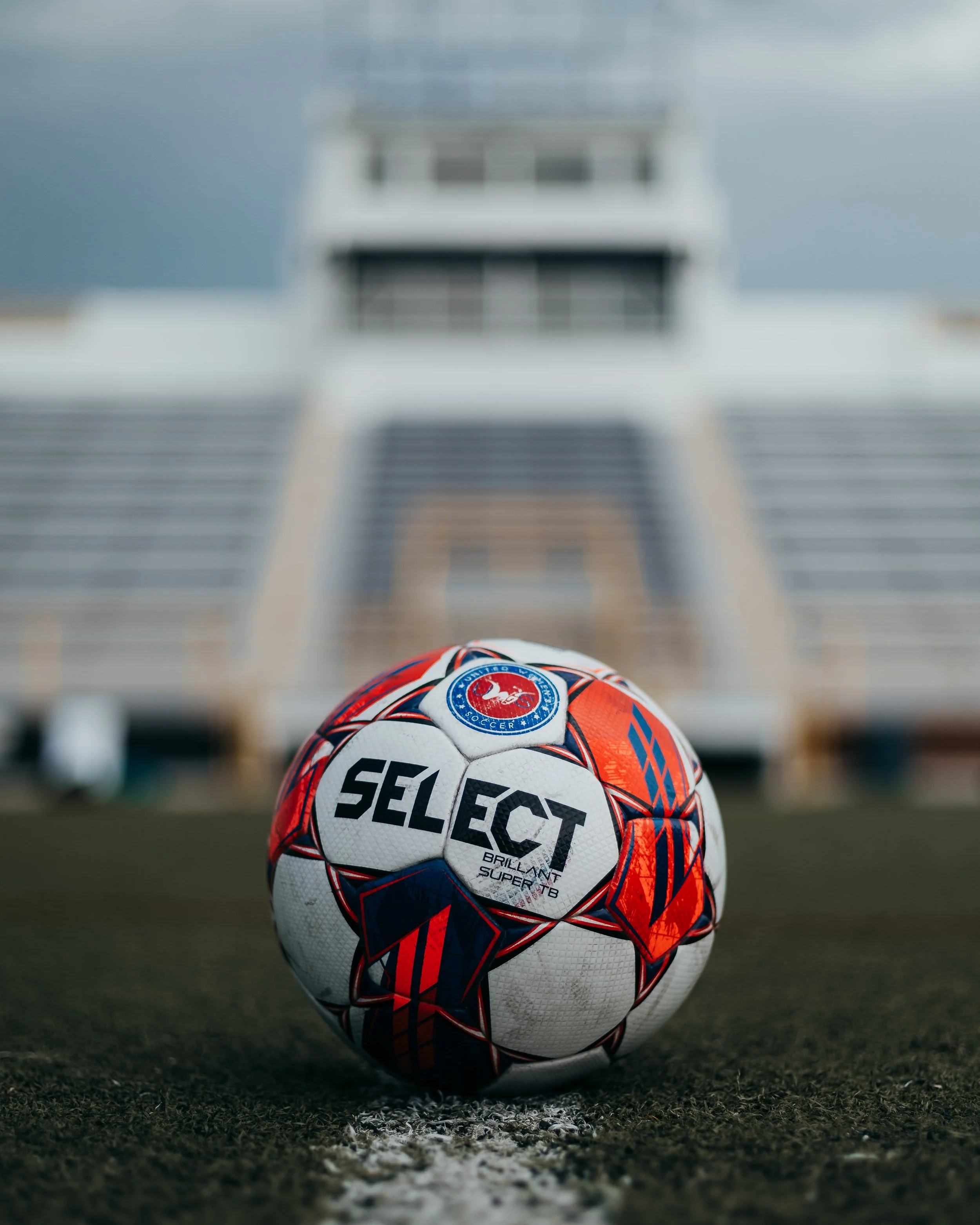 Soccer ball on a field with stadium seats and a press box in the background.