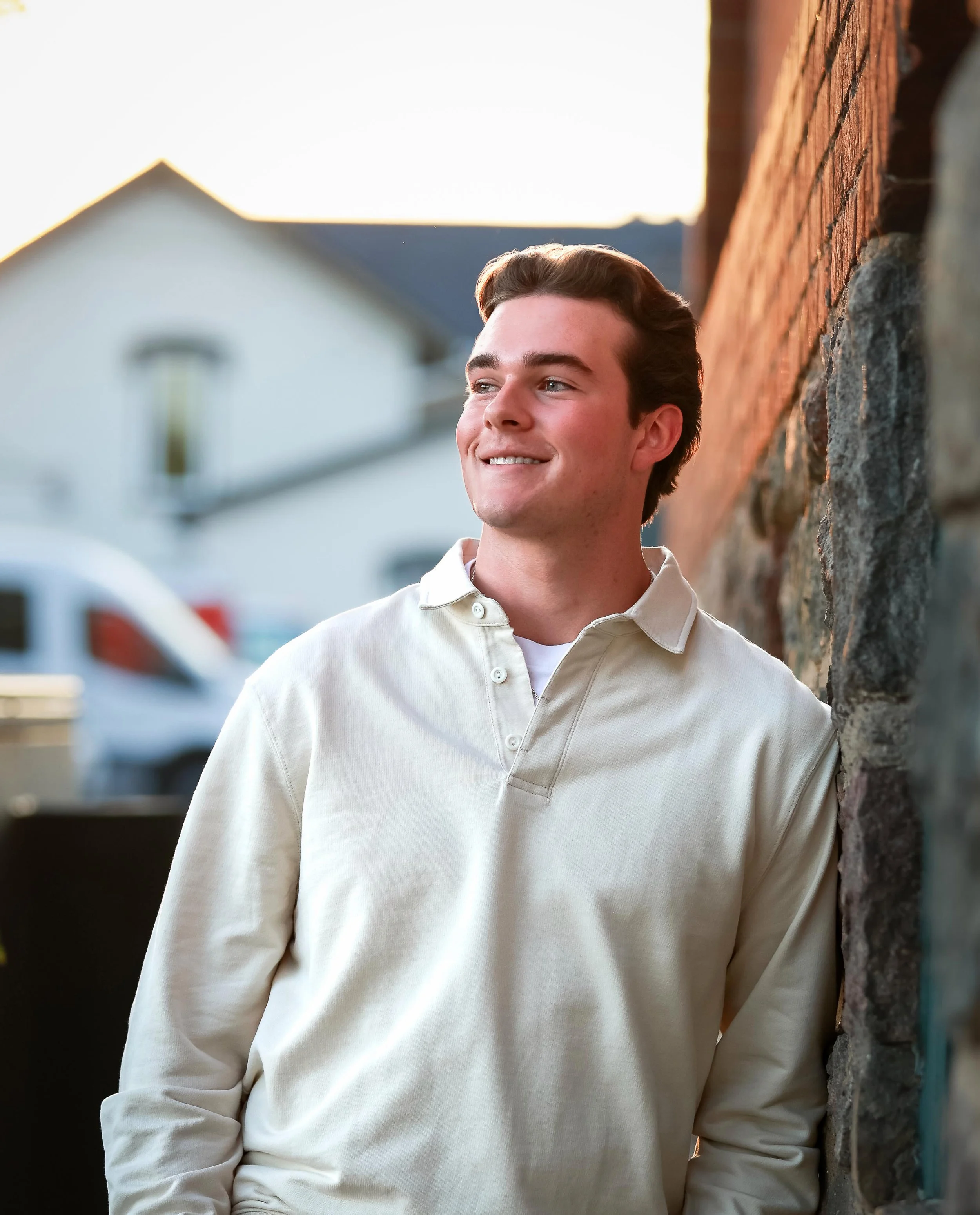 A young man in a cream-colored polo shirt with a slight smile, standing outdoors and leaning against a brick wall during sunset.