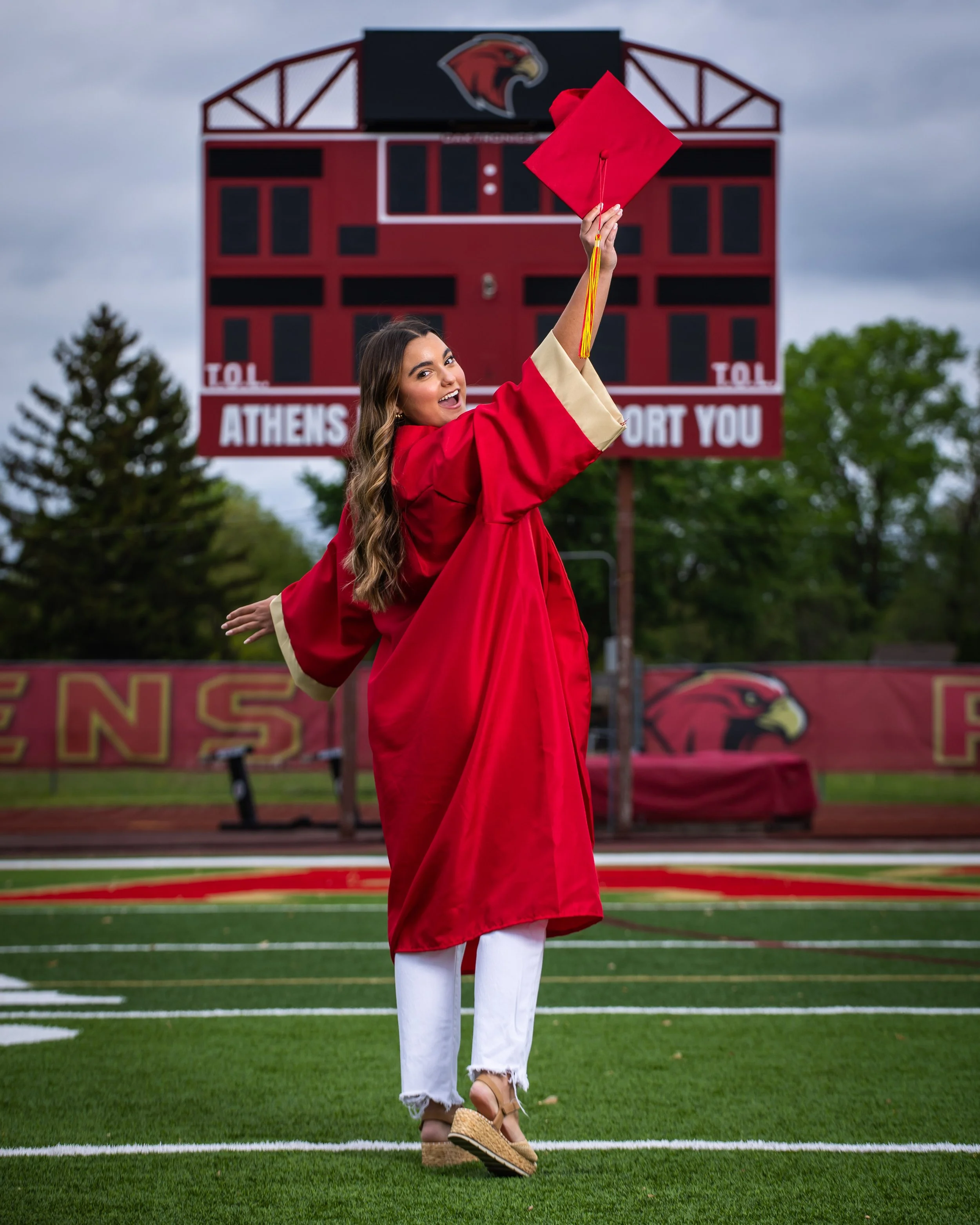 A woman in a red graduation gown and cap stands on a football field, smiling and holding her cap in the air. In the background, there is a scoreboard and a banner with a hawk logo and the words "TOL ATHENS SUPPORT YOU." The woman is wearing white pan