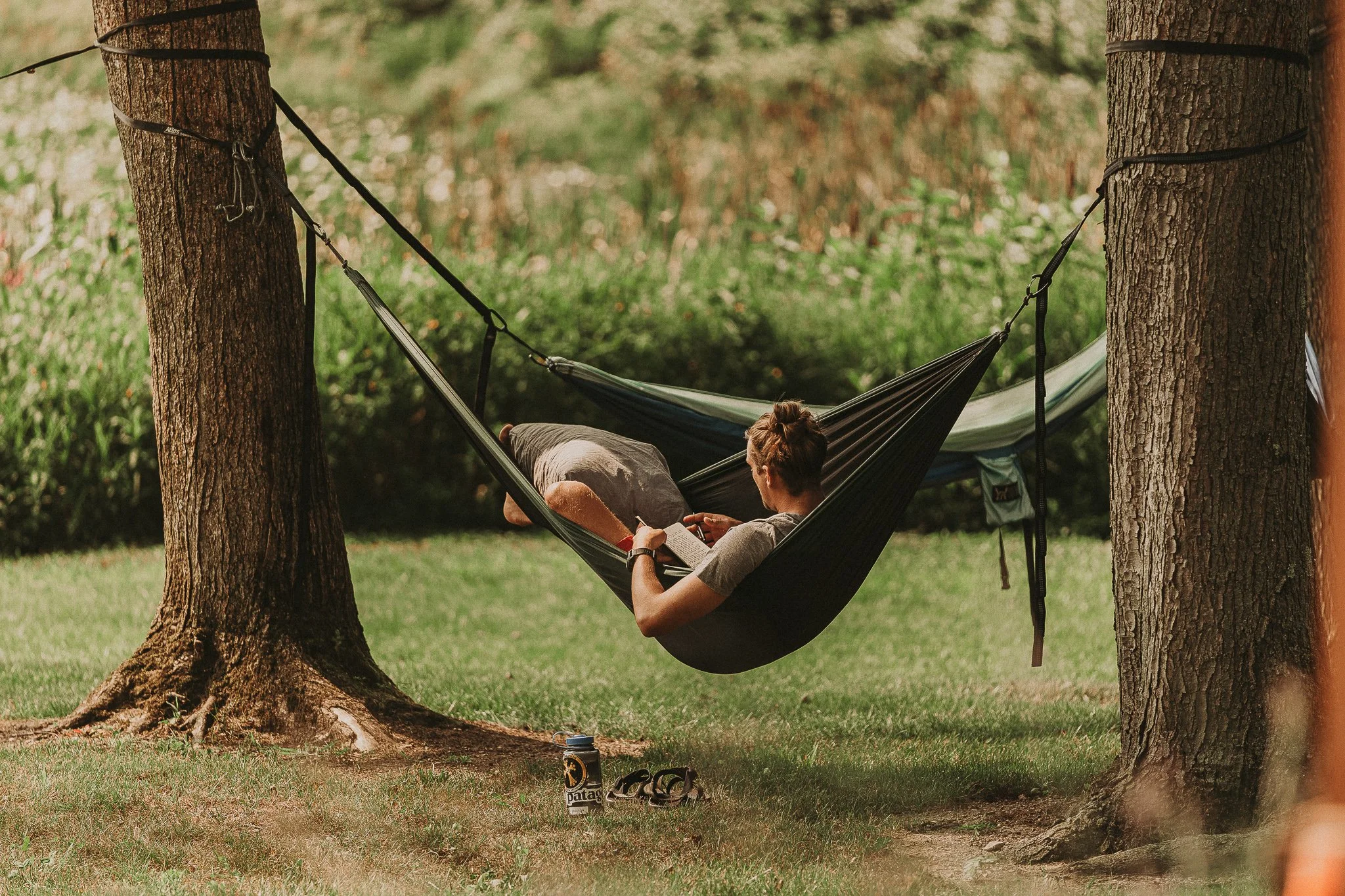 Person lying in a hammock tied between two trees, reading a book, with a Patagonia water bottle and sunglasses on the grass nearby.