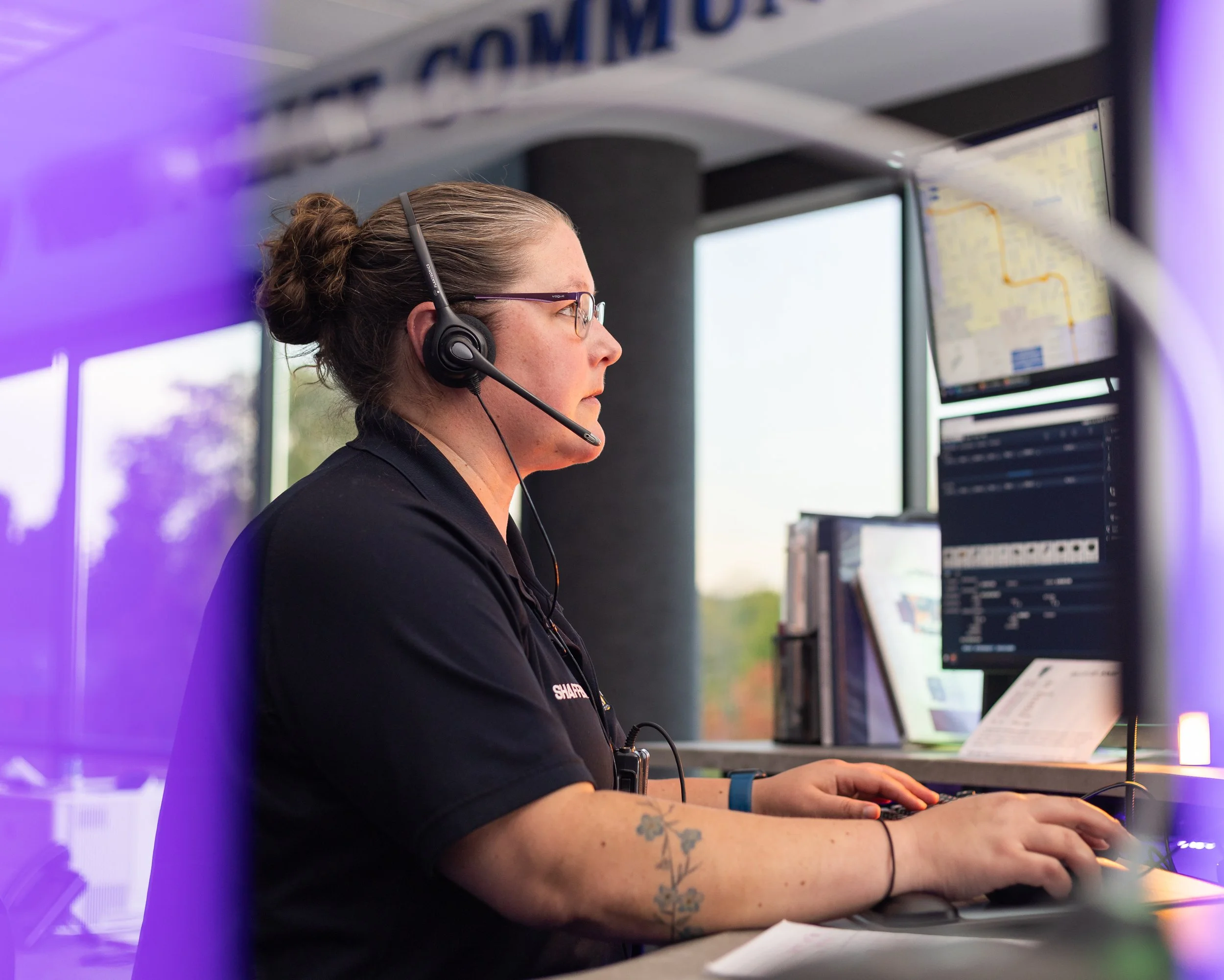 A woman with glasses and a bun wearing a headset sitting at a desk with two computer monitors.