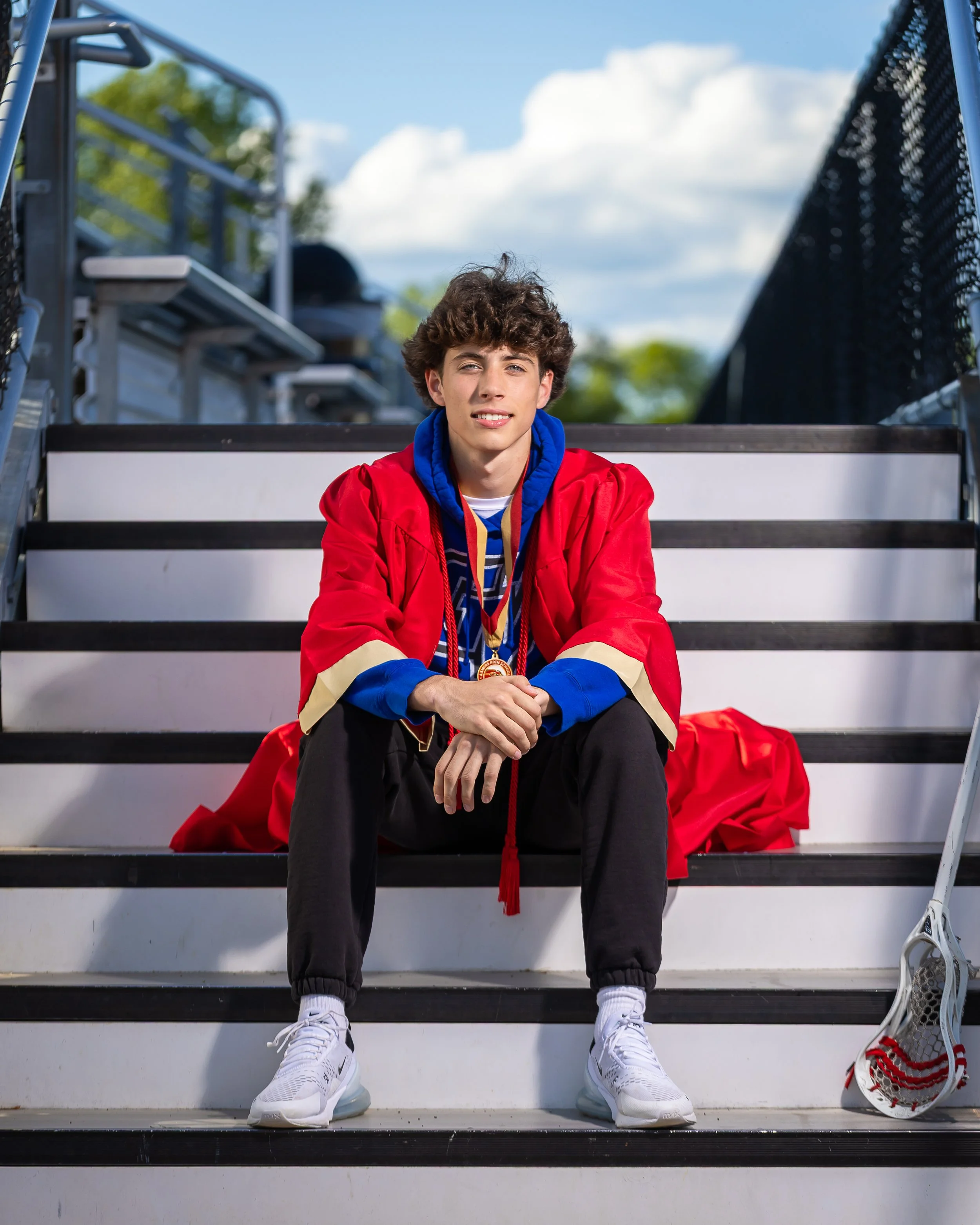 A young man in a red sports jacket with gold and blue accents, sitting on white and black outdoor stairs, holding a medal, with a lacrosse stick next to him, and a clear sky with some clouds in the background.