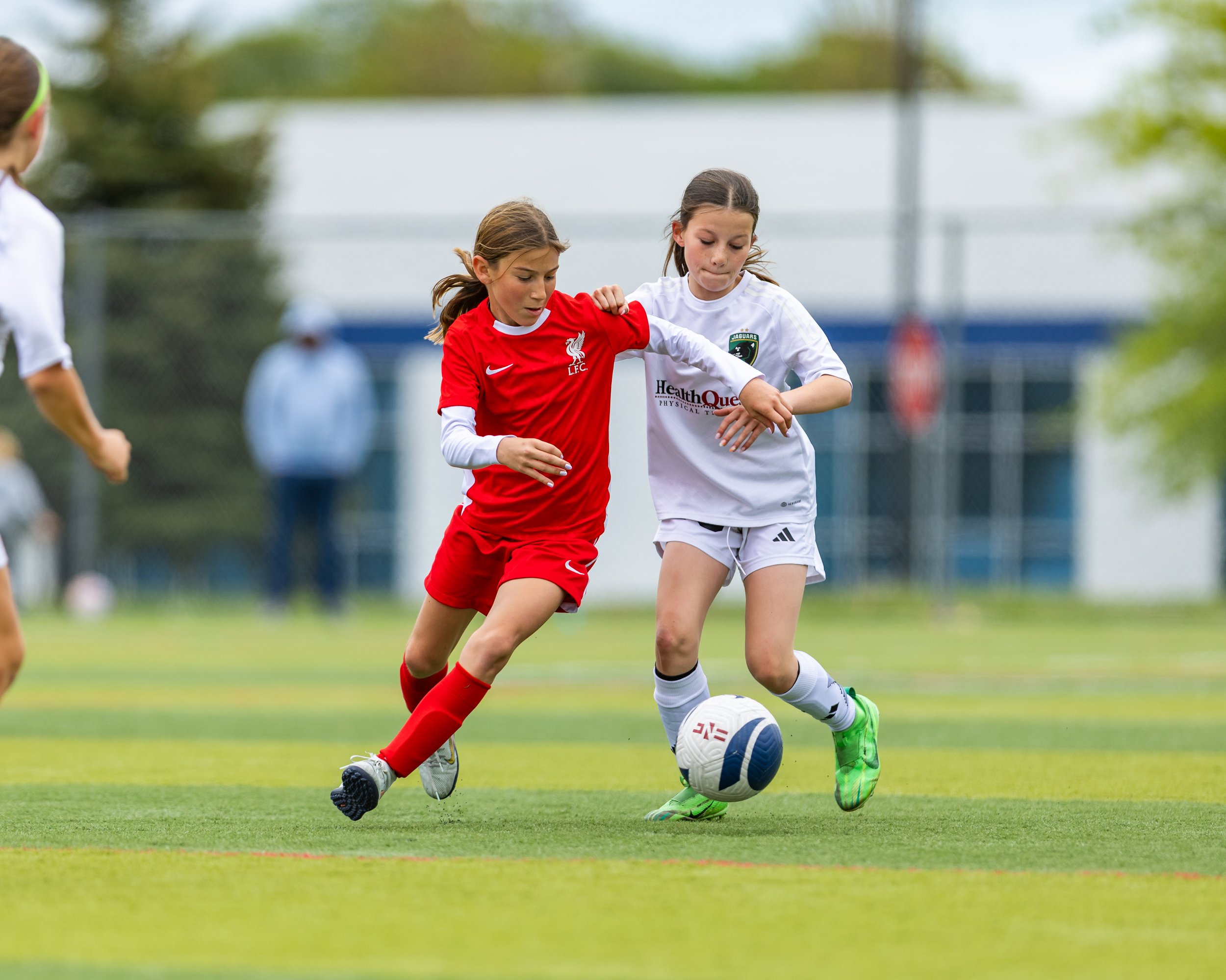 Two young girls playing soccer on a green field, competing for the ball. One girl in a red Liverpool jersey and the other in a white jersey with green shoes.