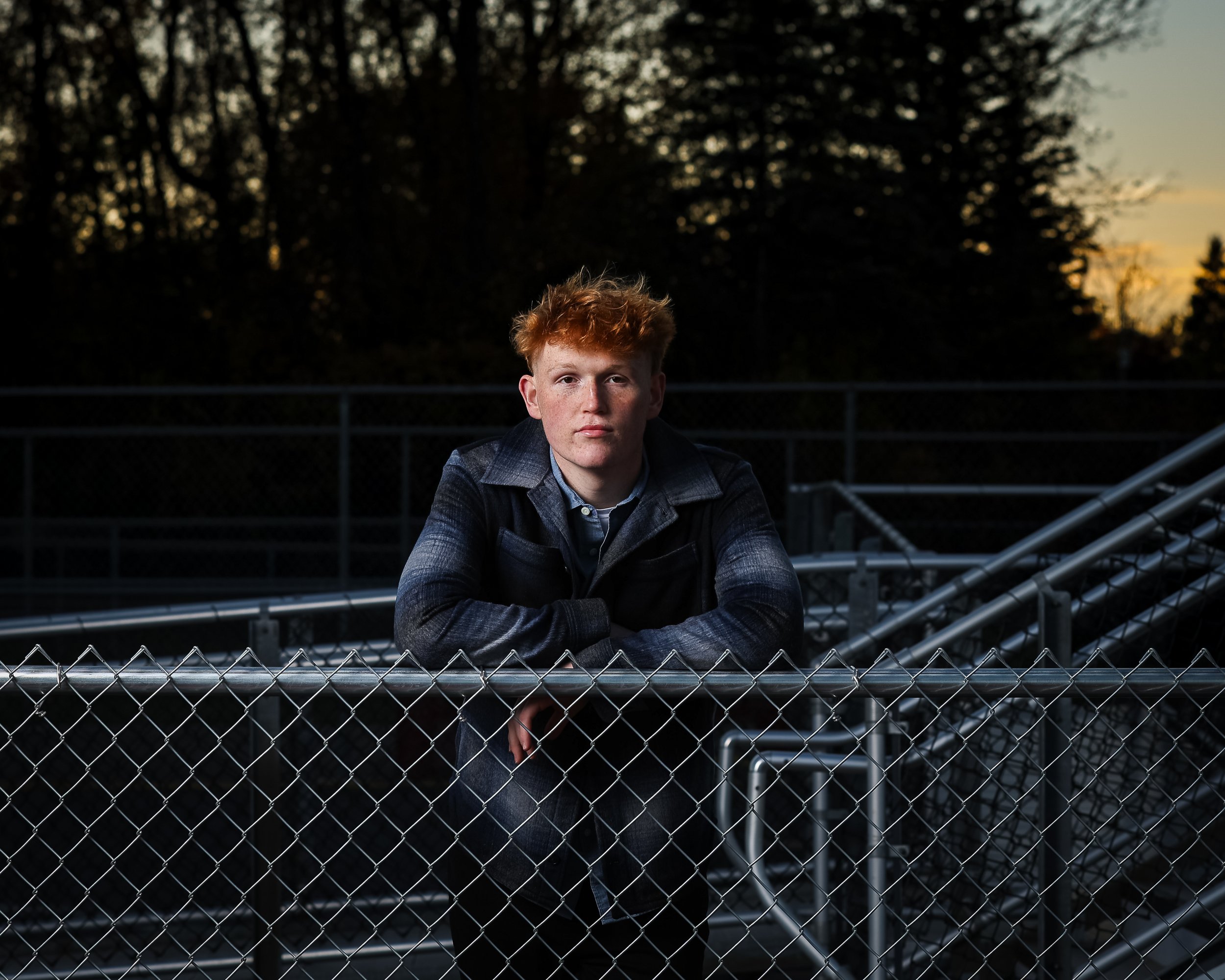 A young man with red hair and fair skin leaning on a chain-link fence, outdoors at dusk, with trees in the background and a sunset sky.