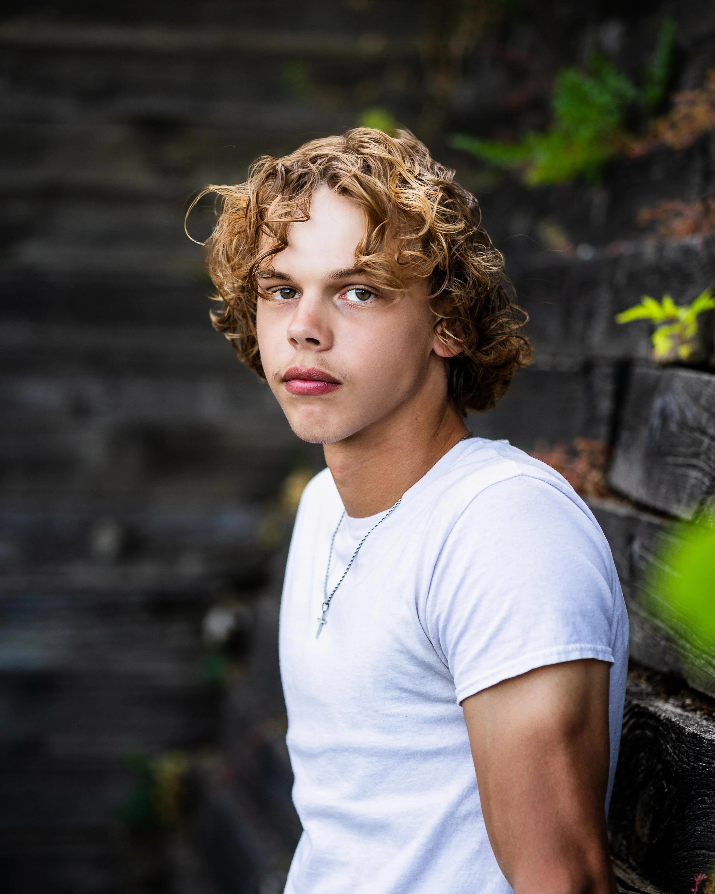 A young man with curly blond hair and blue eyes wearing a white t-shirt and a necklace with a cross, leaning against a wooden wall outdoors.