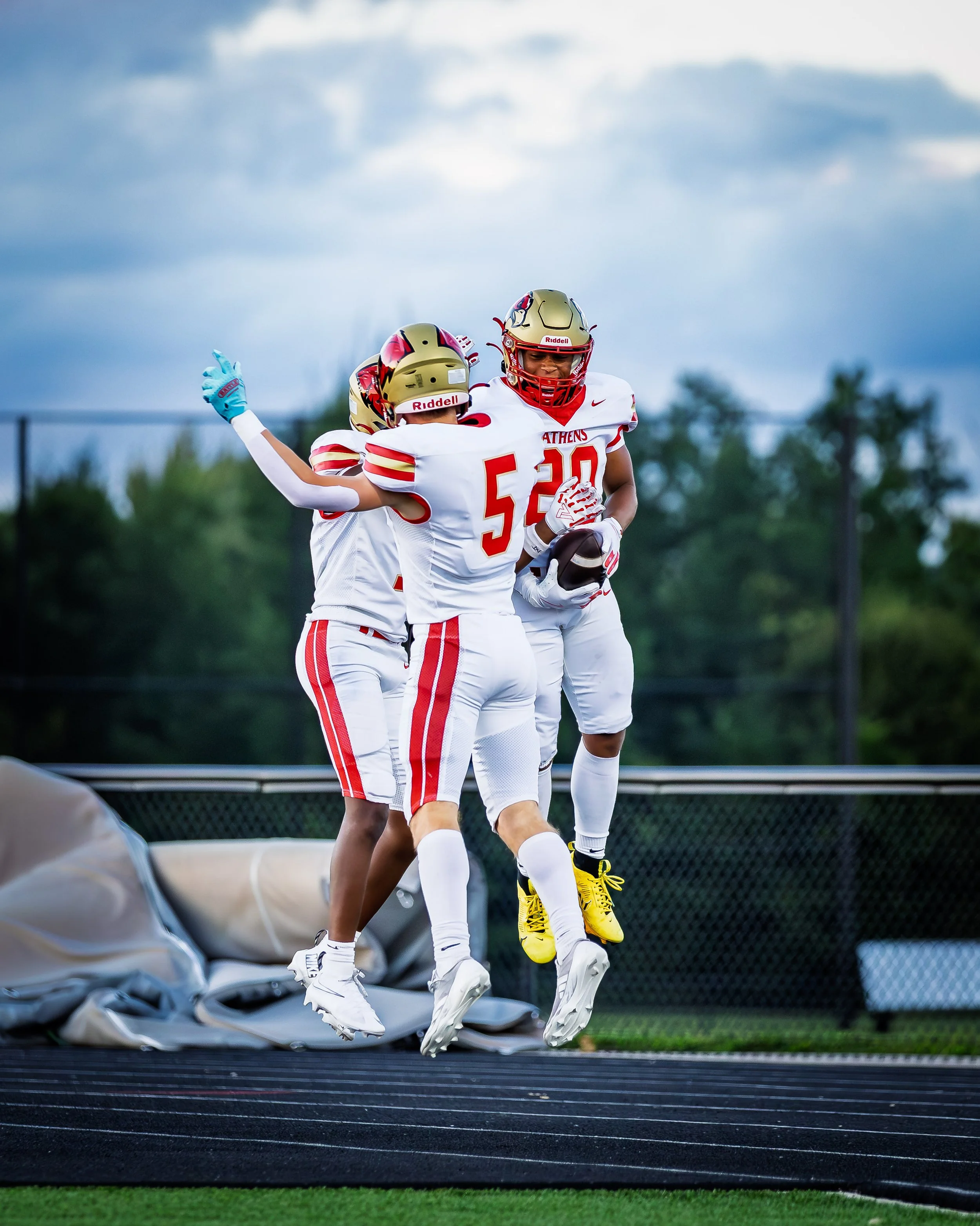 American football players in white and red uniforms celebrating on the field, with one player jumping and holding a football, under a cloudy sky.
