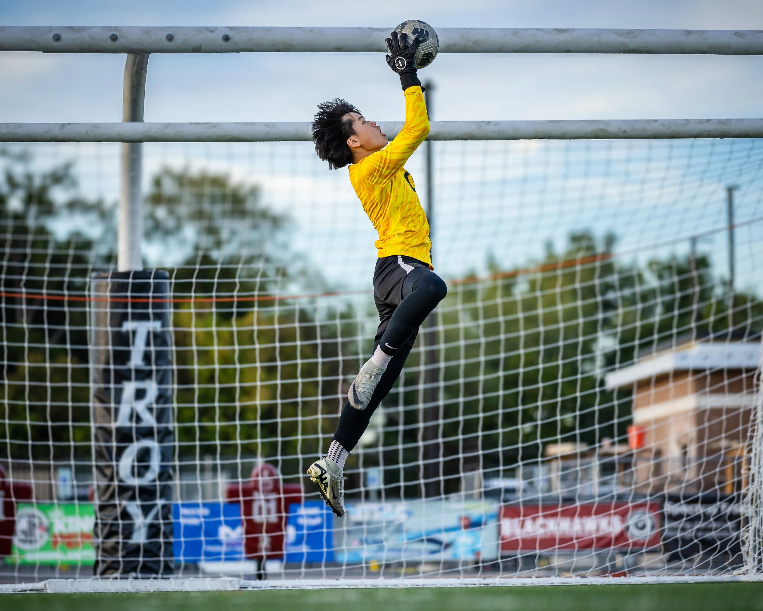 Soccer goalkeeper in yellow jersey, black shorts, and grey gloves jumping to catch a ball in front of the goal during a match.