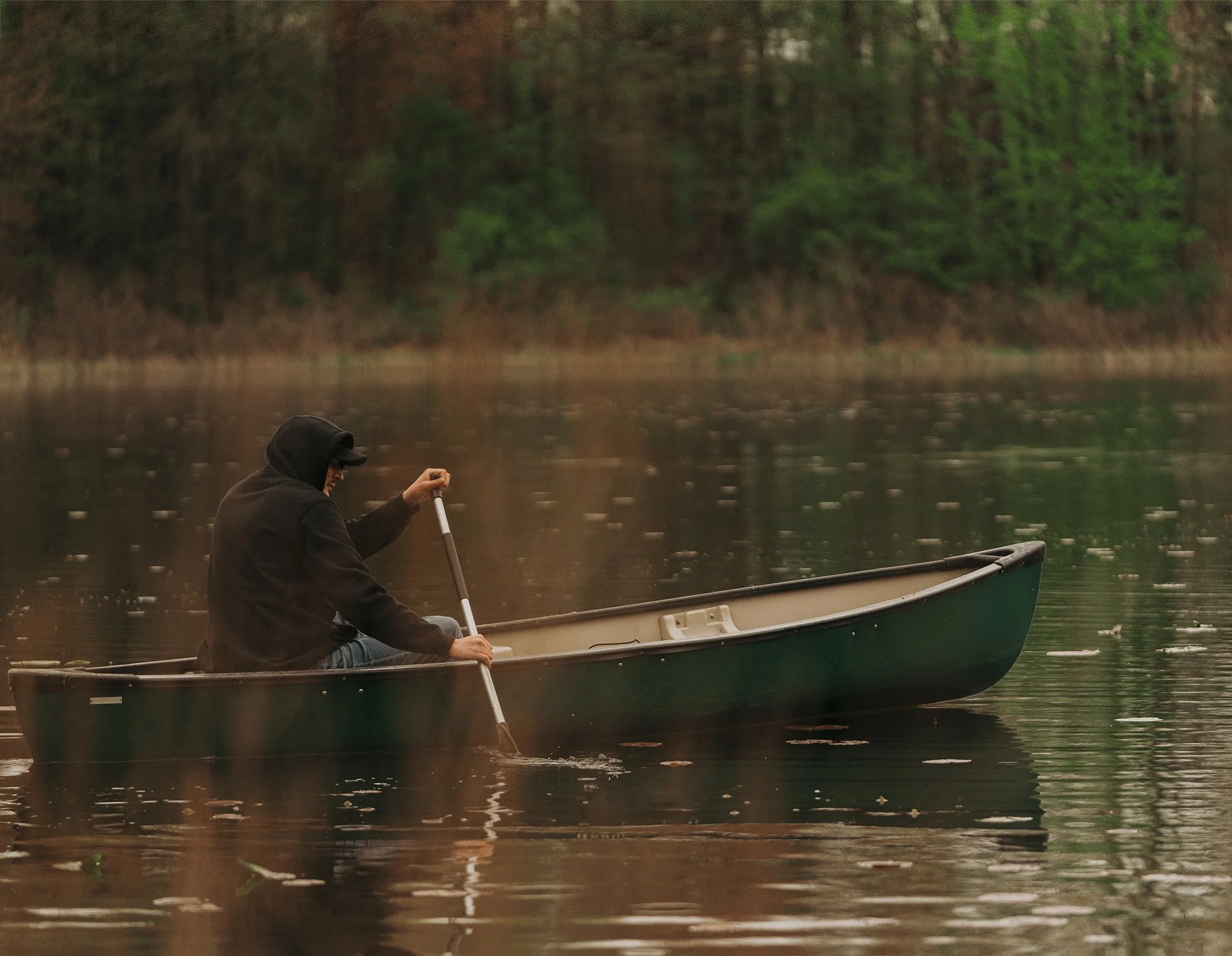 A person wearing a black hoodie and cap paddling a canoe on a calm lake with a forested shoreline in the background.