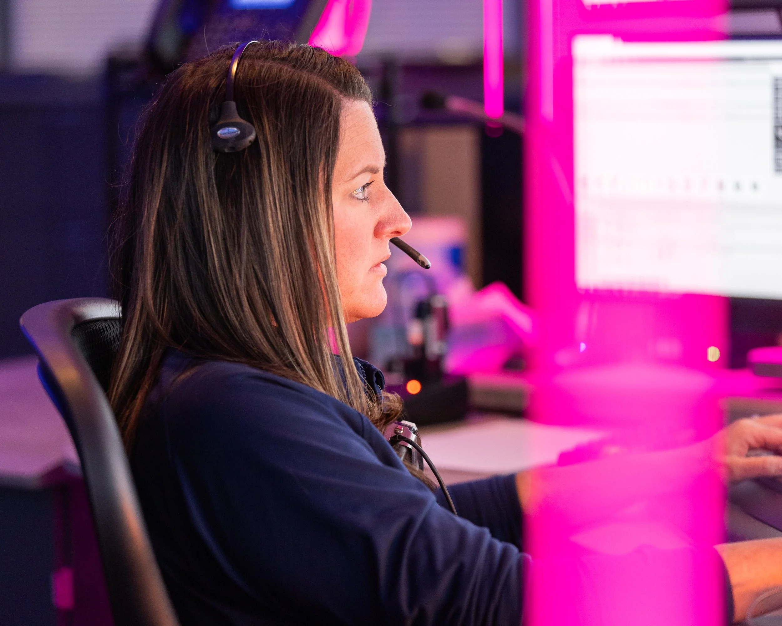 A woman with long brown hair wearing a headset with microphone, sitting at a computer desk in a neon-lit office