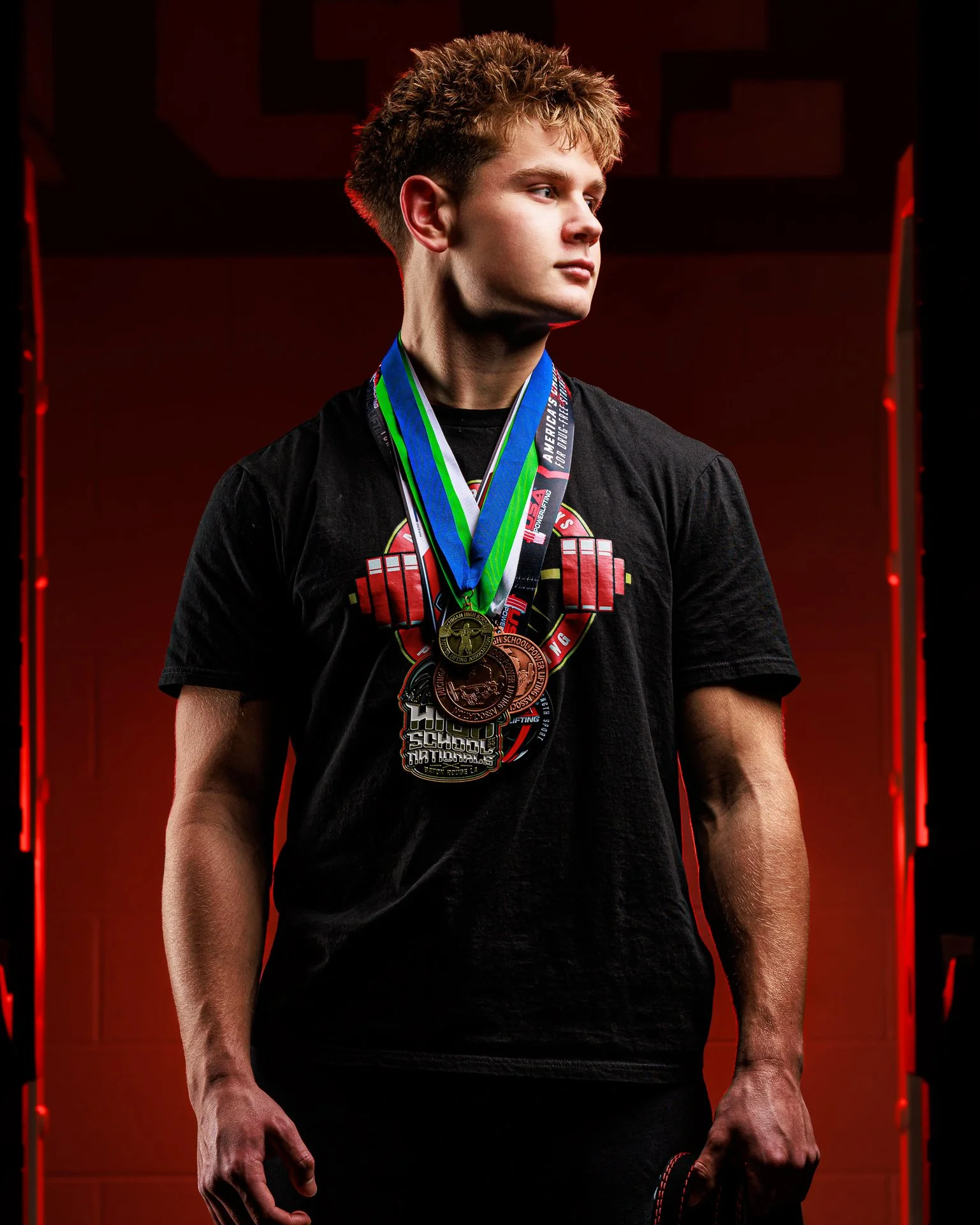 Young male athlete with medals around his neck, wearing a black t-shirt with a weightlifting graphic, looking to the side, posed against a dark red background with red accents.