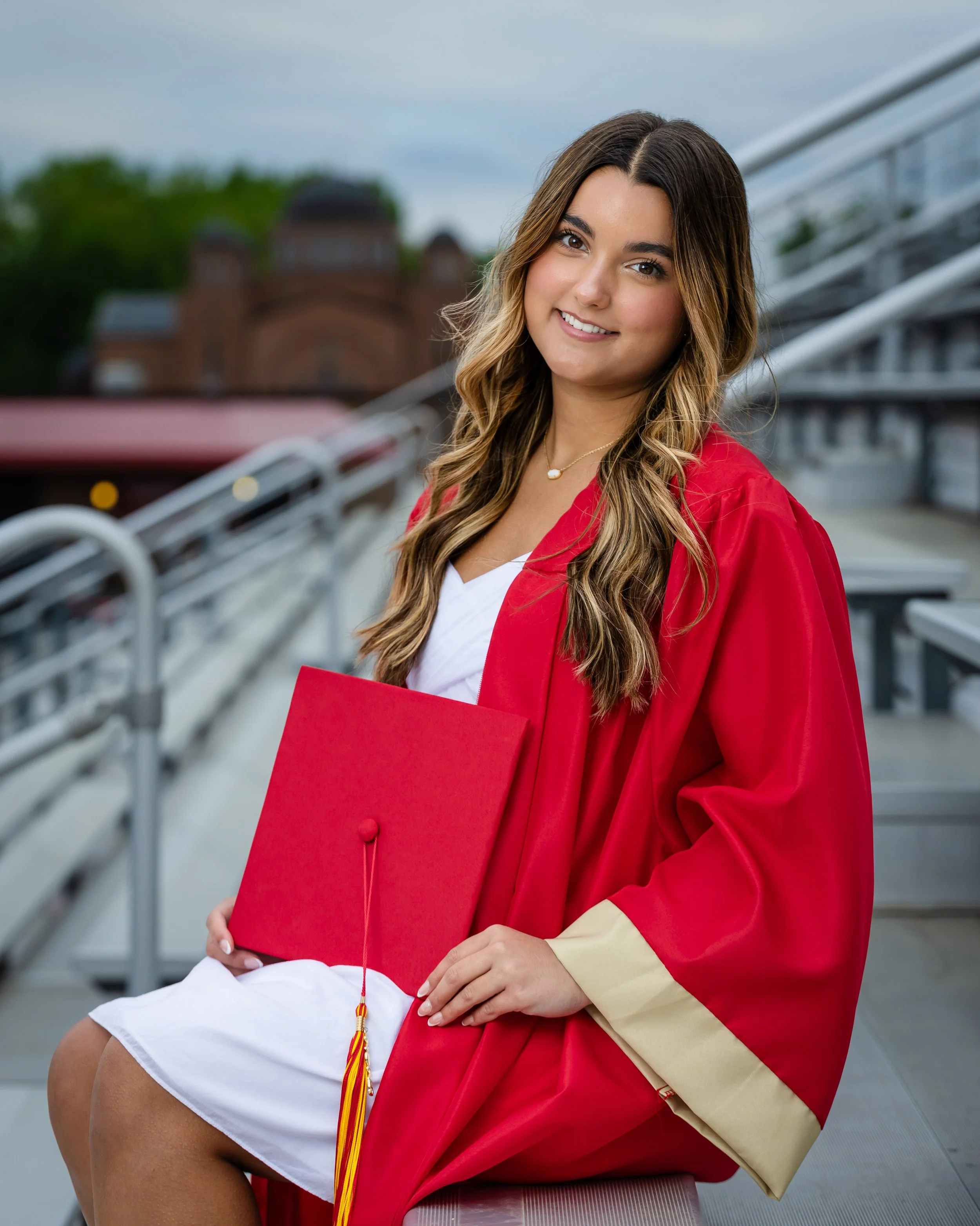 Young woman in a red graduation gown sitting on a bench, holding a red diploma cover, with stadium seats and a historic building in the background.