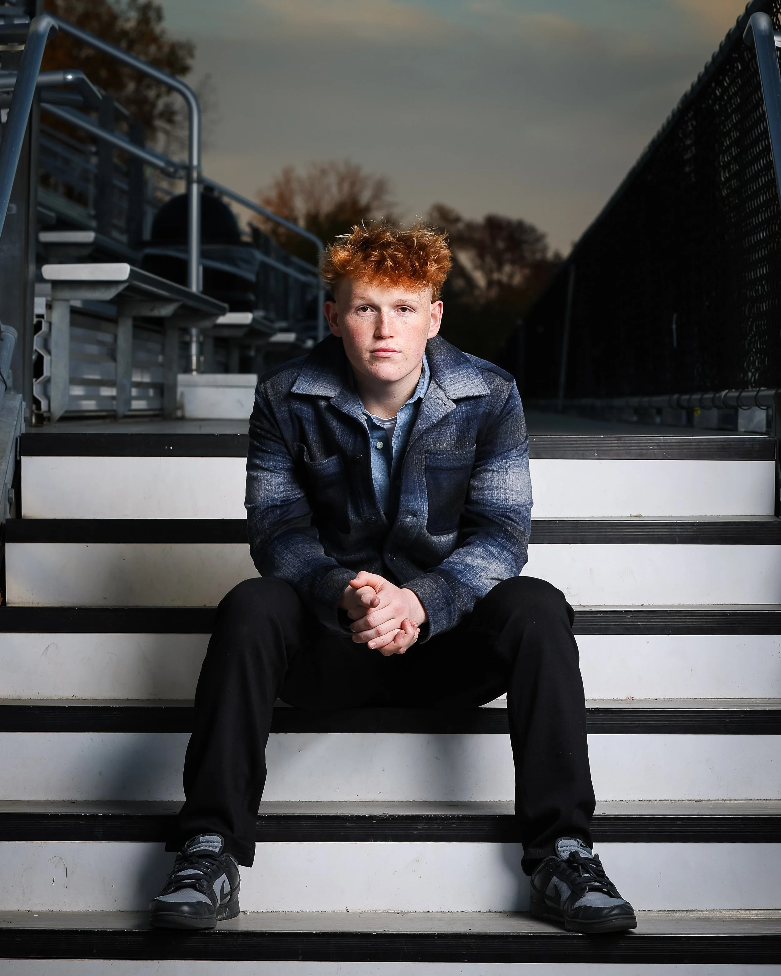 A young man with red hair sitting on outdoor stadium bleachers at sunset, wearing a dark denim jacket, gray shirt, black pants, and sneakers.