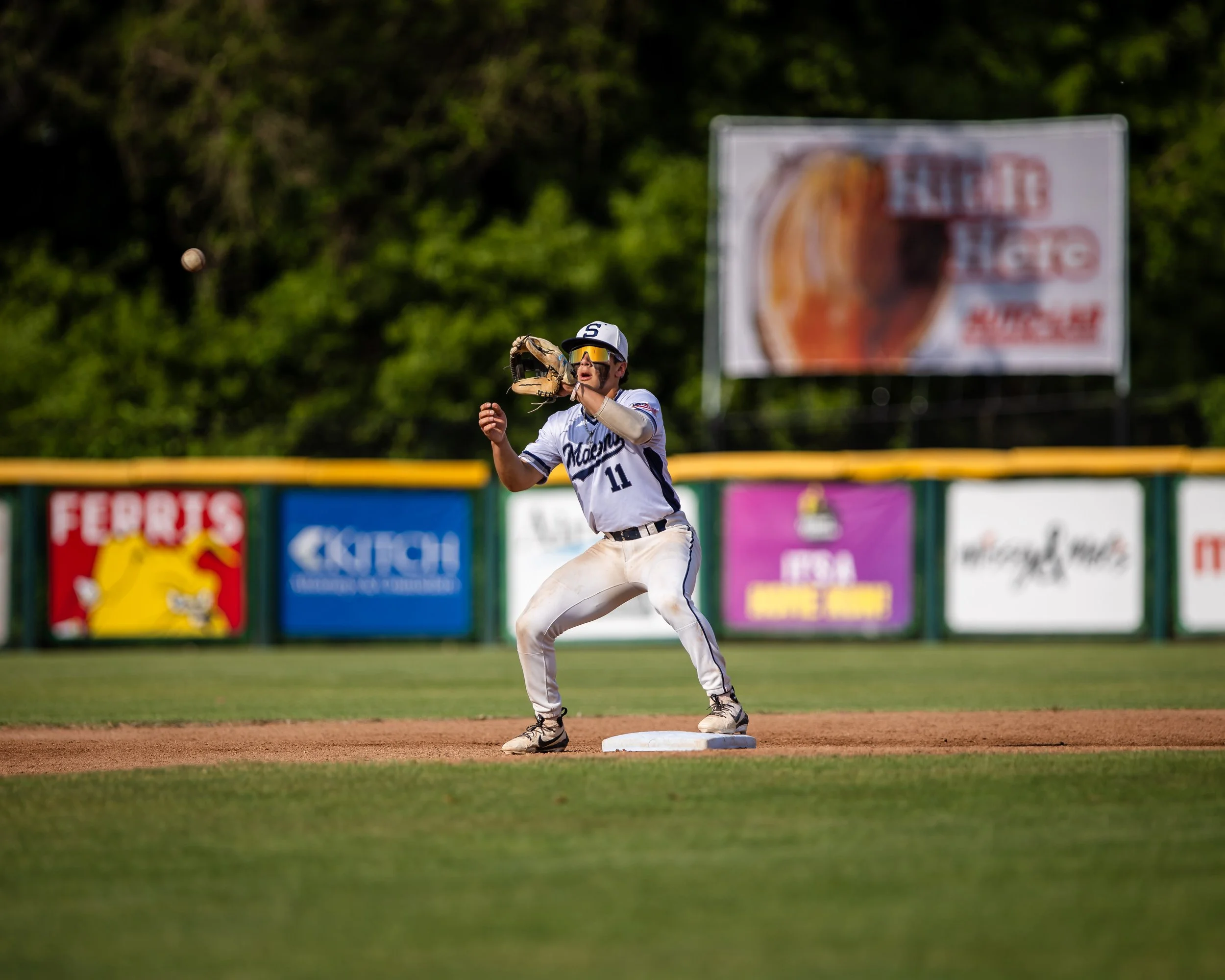 A baseball player in a white uniform with the number 11, standing on the base path, preparing to catch a ball in his glove during a game.