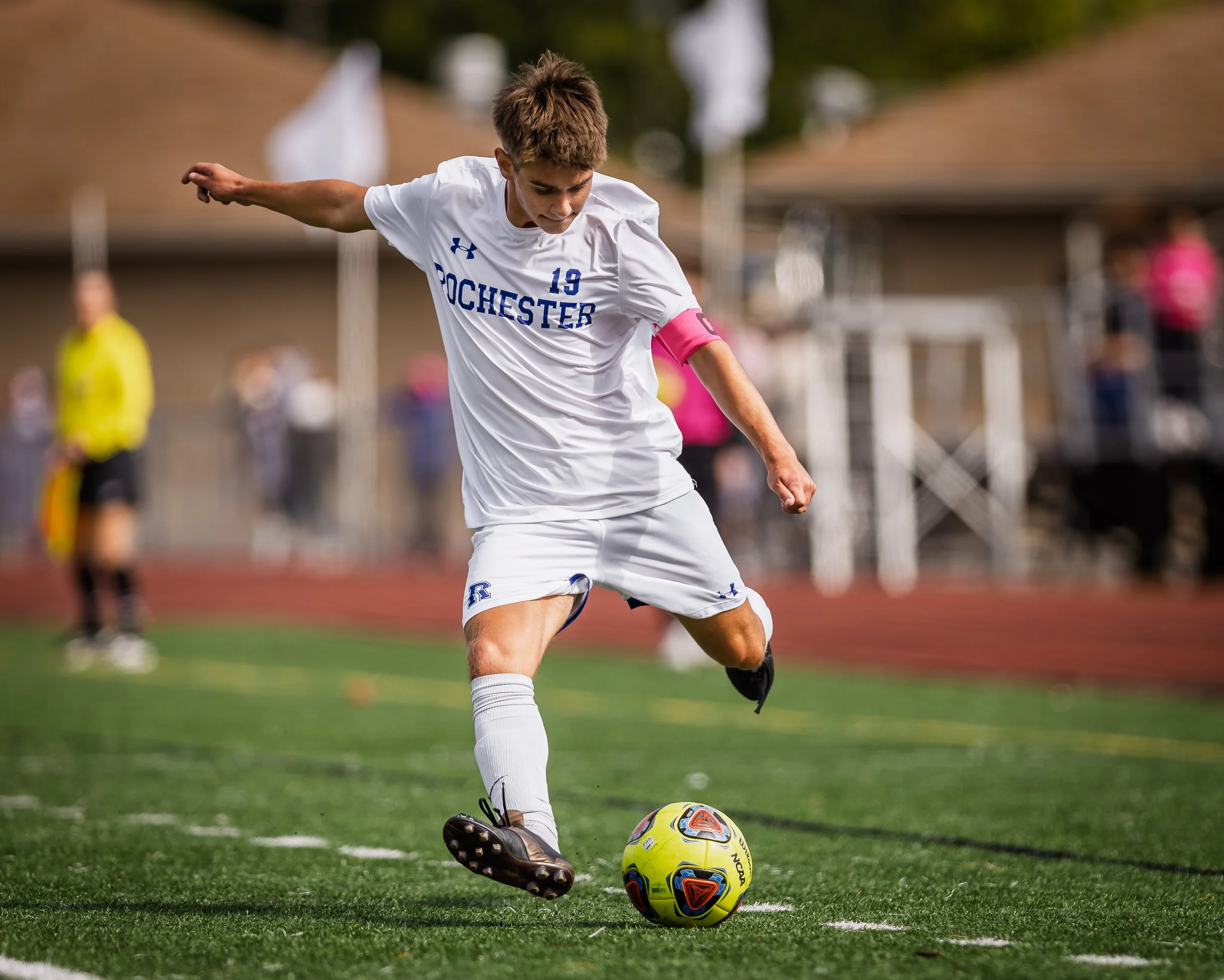 Young male soccer player in white uniform kicking a soccer ball on a field during a game.