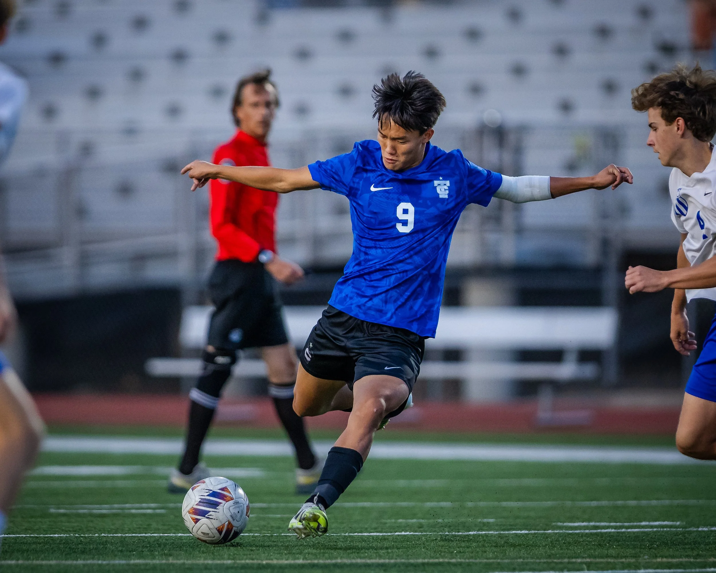 A young male soccer player in a blue jersey with the number 9 kicks a soccer ball on a field during a game.