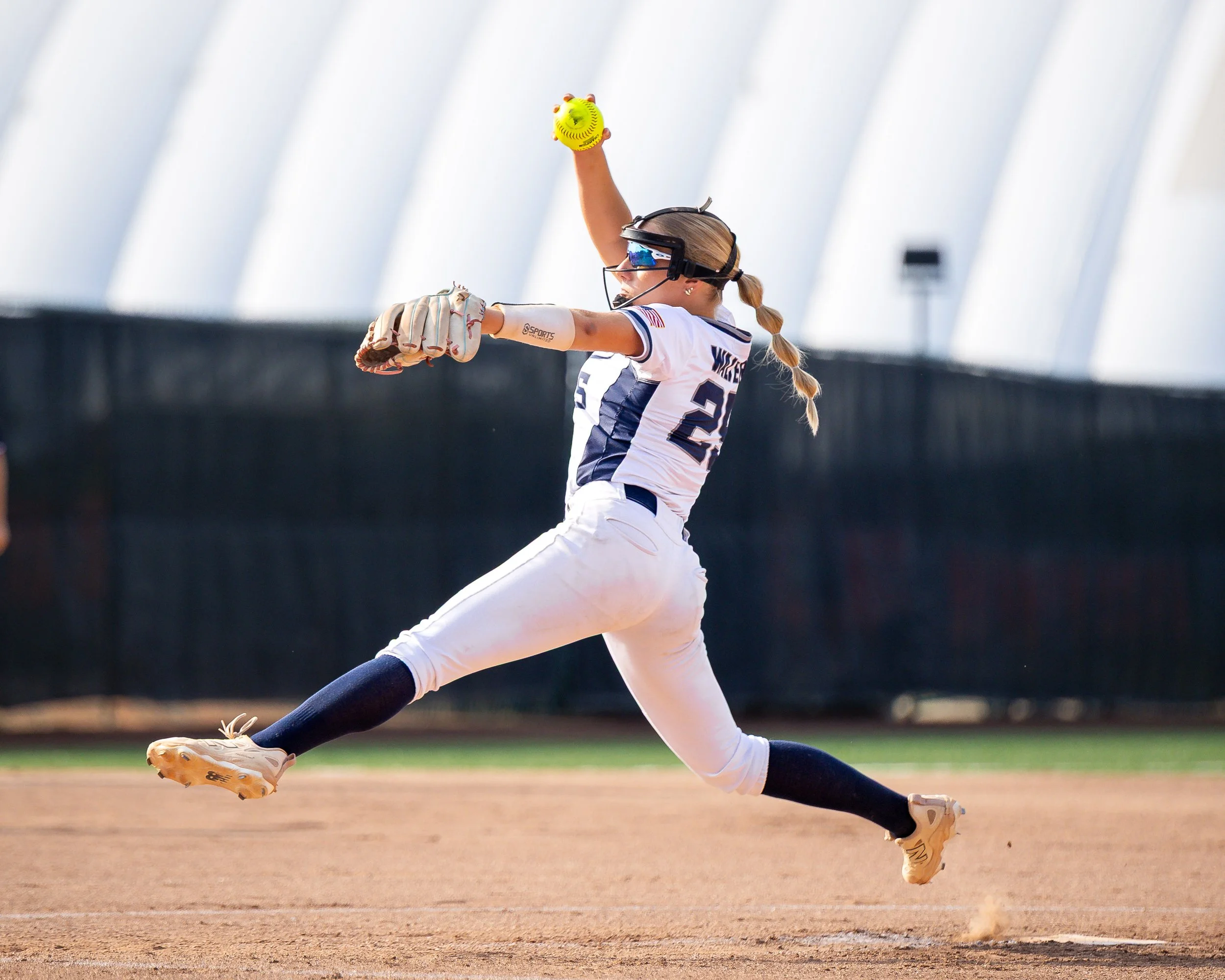 Softball player in white uniform with navy accents diving to catch a ball at second base, wearing goggles, gloves, and a braid in her hair.