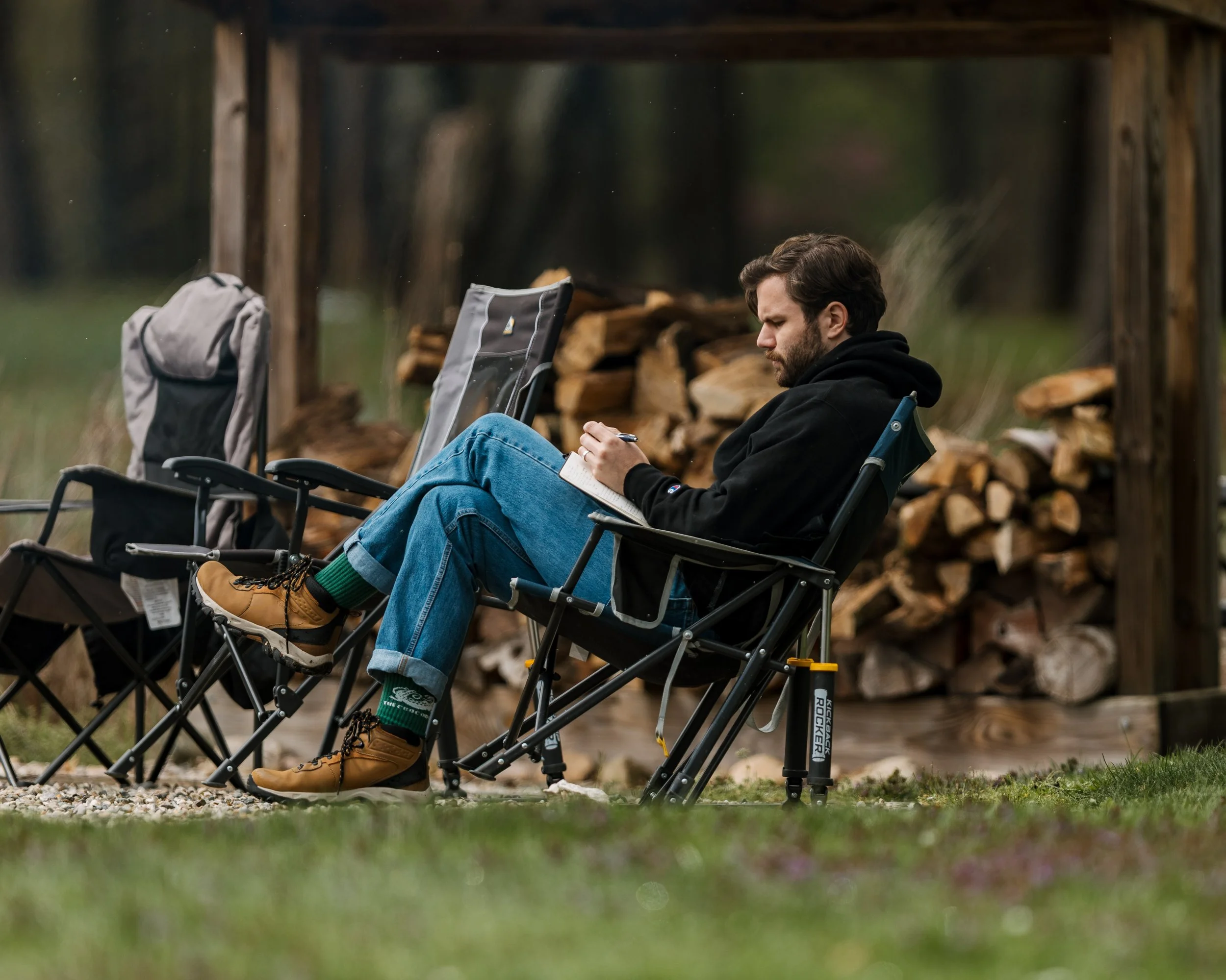 A man sitting in a camping chair outdoors, writing in a notebook with stacked firewood in the background.