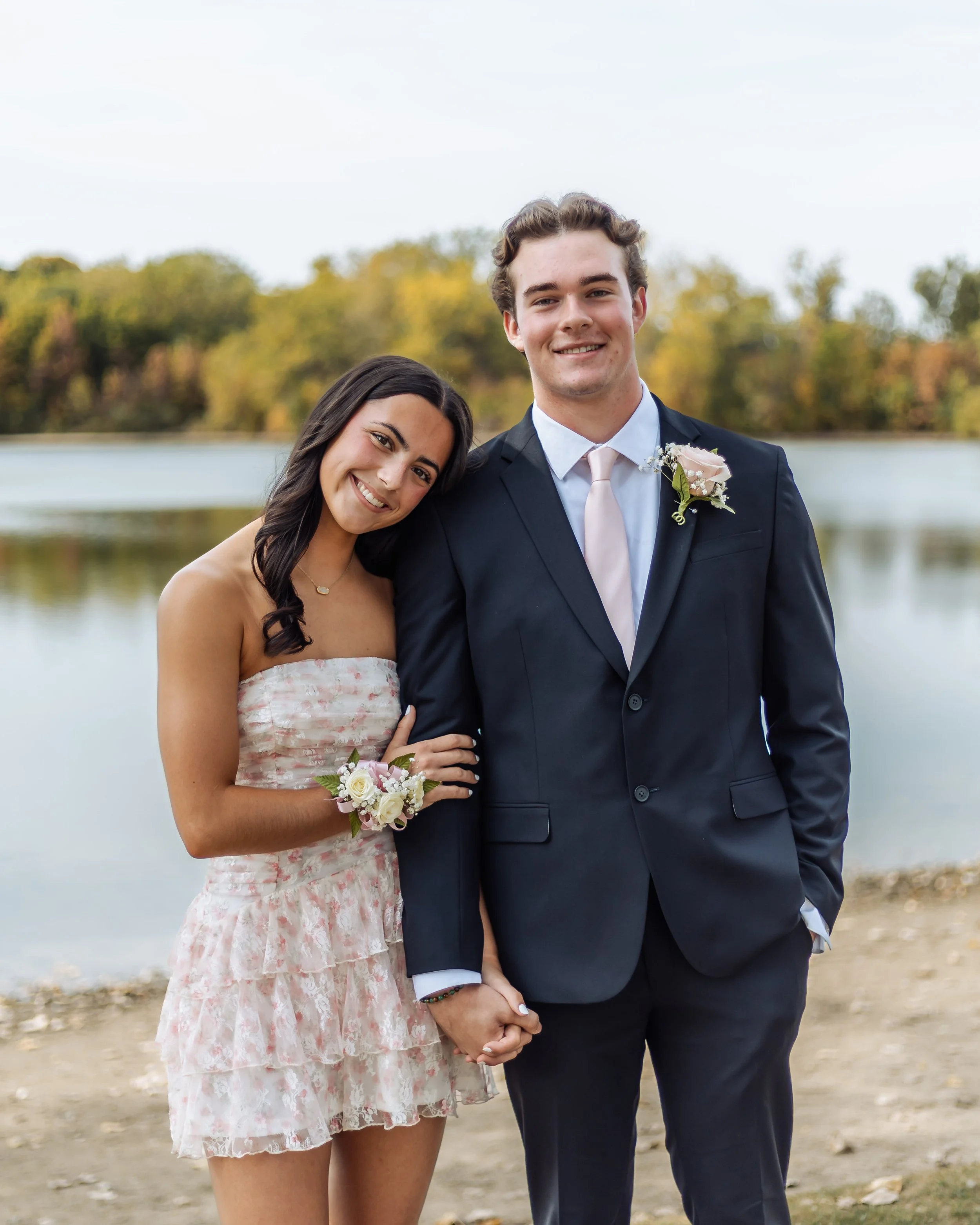 A young couple dressed in wedding attire, standing outdoors near a body of water, with trees displaying fall foliage in the background. The woman is leaning her head on the man's shoulder and holding his hand, both smiling.