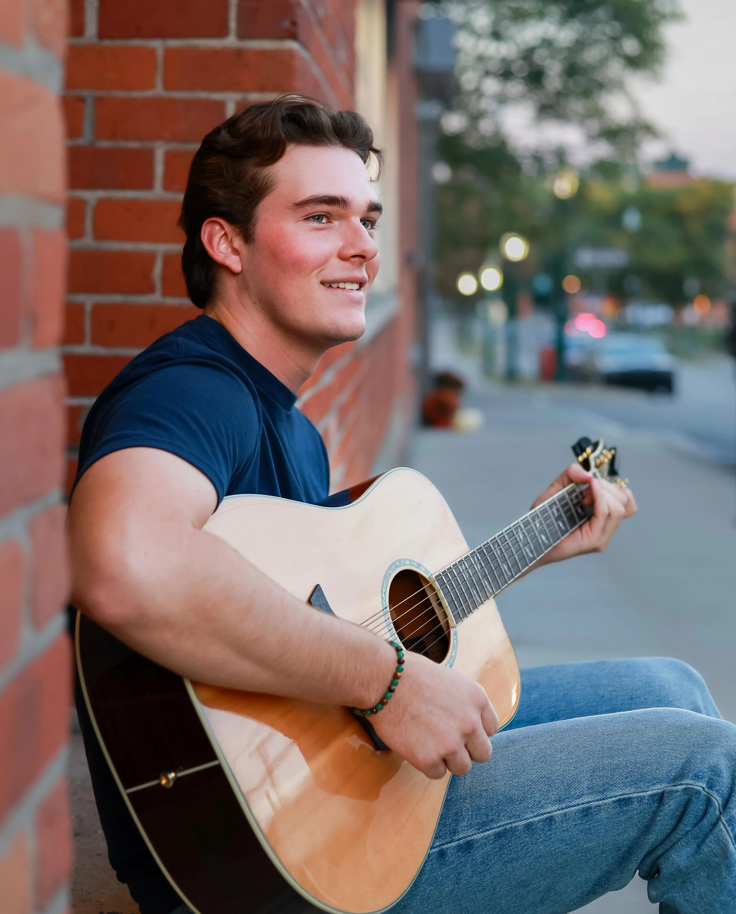 Young man playing guitar and sitting against a brick wall on a city sidewalk at dusk.